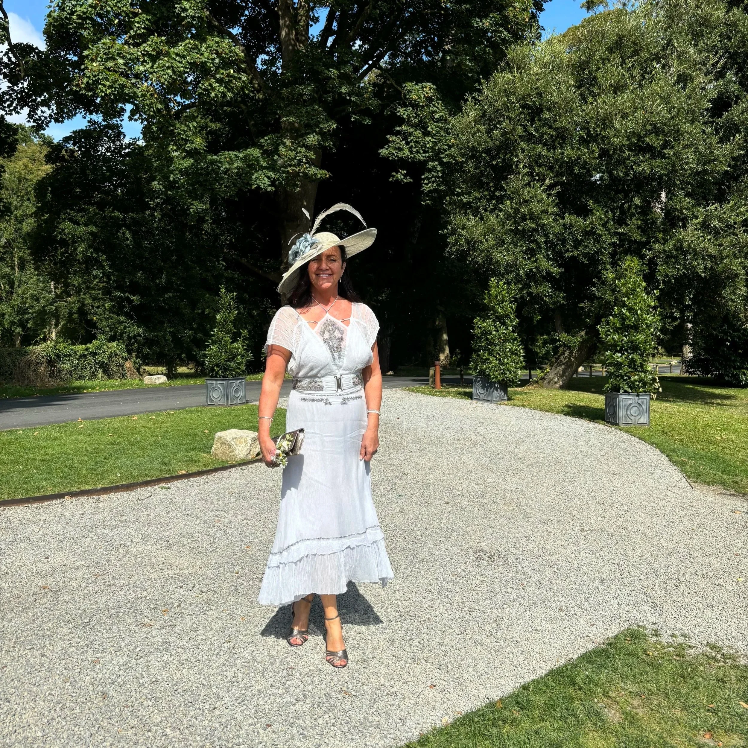 A smiling woman in a white dress and a large, decorative sun hat stands outside in a park with green trees and a paved pathway.