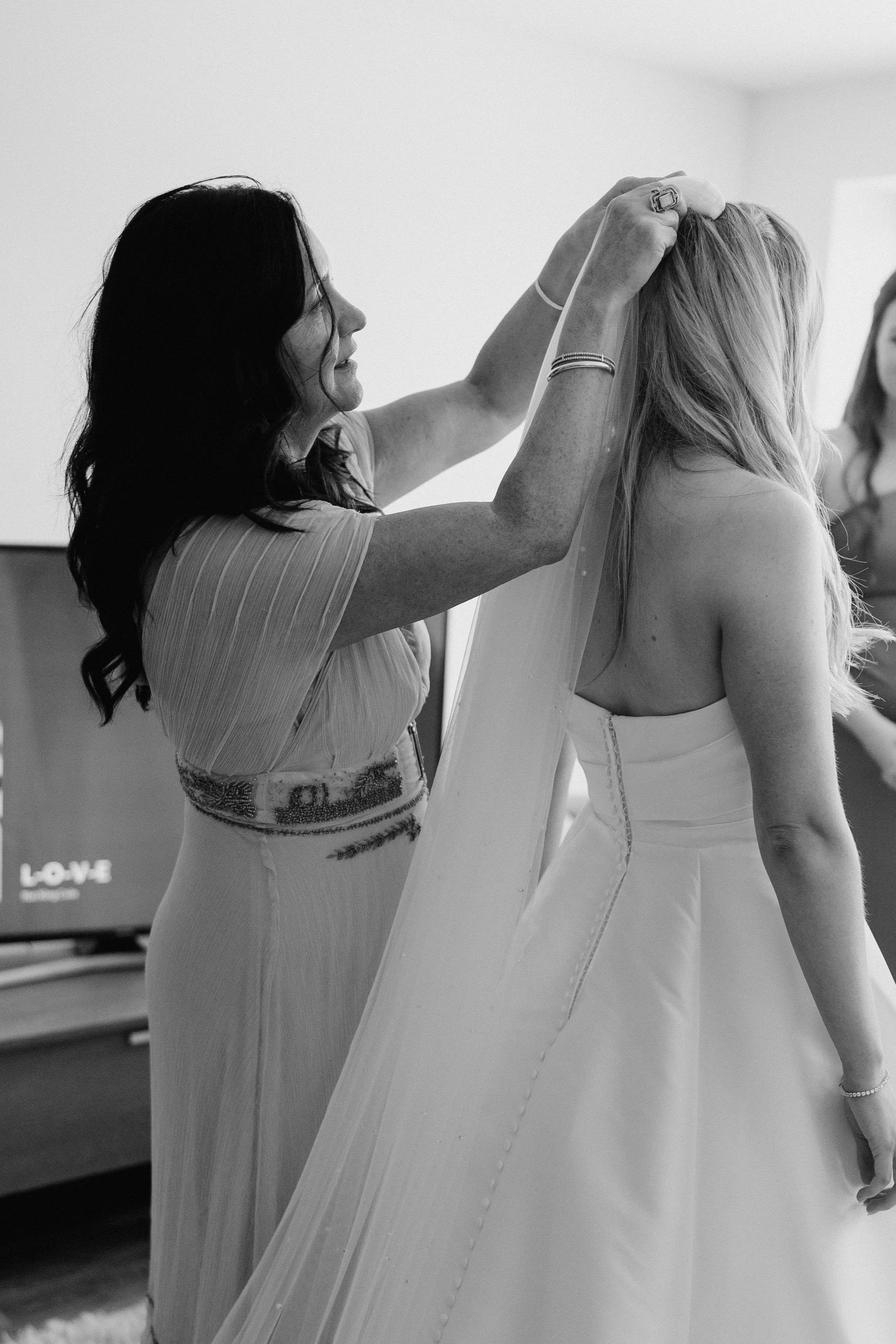 A woman helping another woman with blonde hair adjust her wedding veil in a room with minimal background.