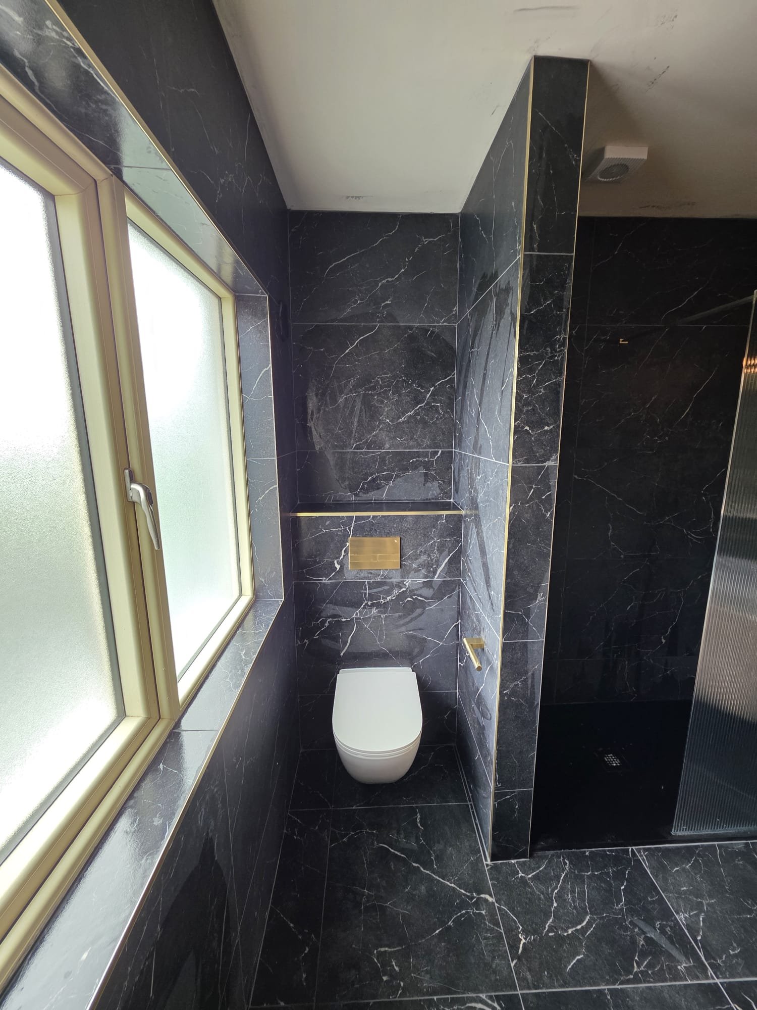 A modern bathroom with black marble walls and floor, a window with frosted glass, a white toilet, and a gold vent on the wall.