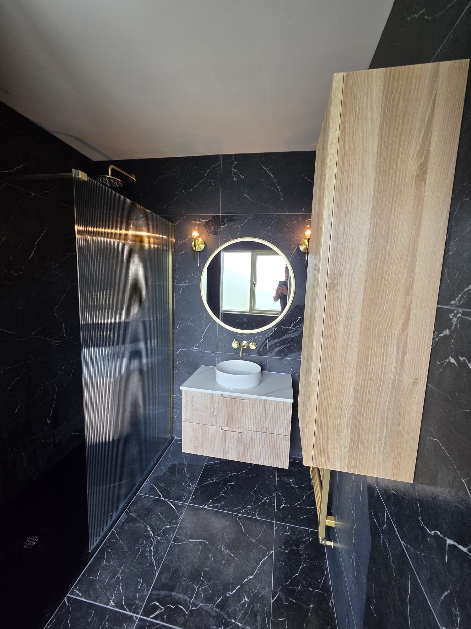 Modern bathroom with black marble walls and floor, a round mirror with backlit LED lighting, two gold wall sconces, a white vessel sink on a wooden vanity, a wood cabinet, and a walk-in shower with a ribbed metal door and a gold showerhead.