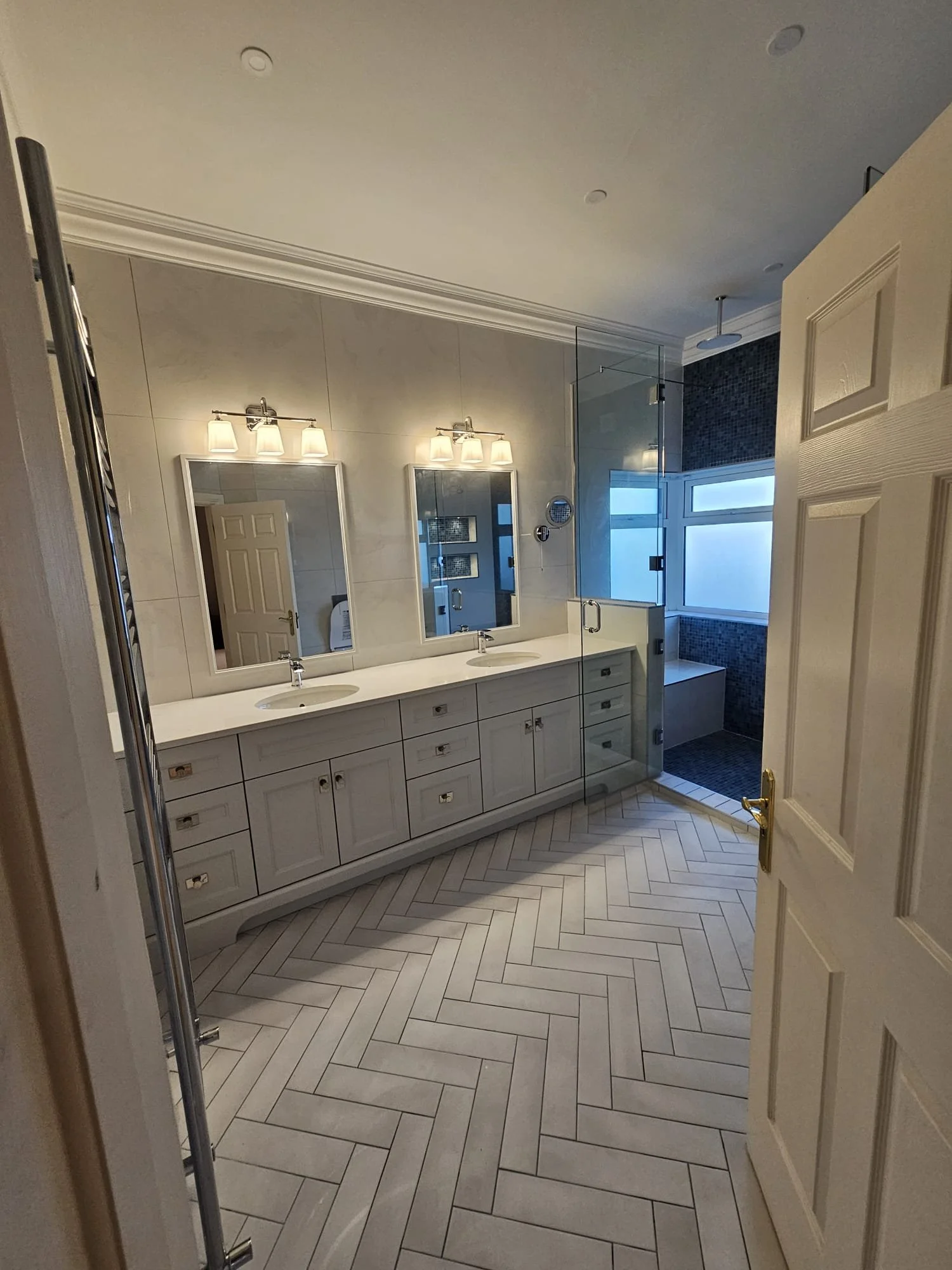 A bathroom with dual mirrors, white cabinetry, and two sinks, illuminated by two light fixtures. There is a shower area with glass doors, opened to a window and a blue-tiled wall.
