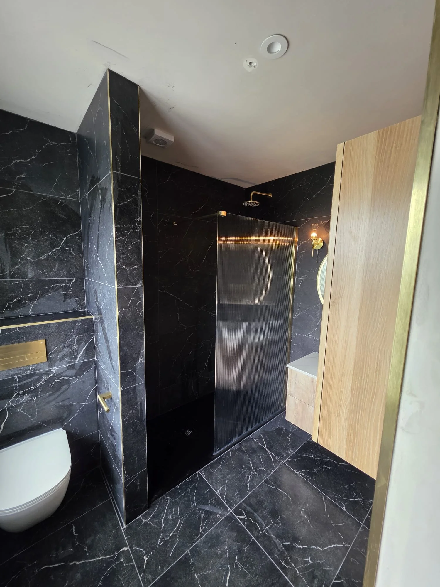 Modern bathroom with black marble walls and floor, a white toilet, a shower area with a rain shower head, a wood cabinet, and gold fixtures.