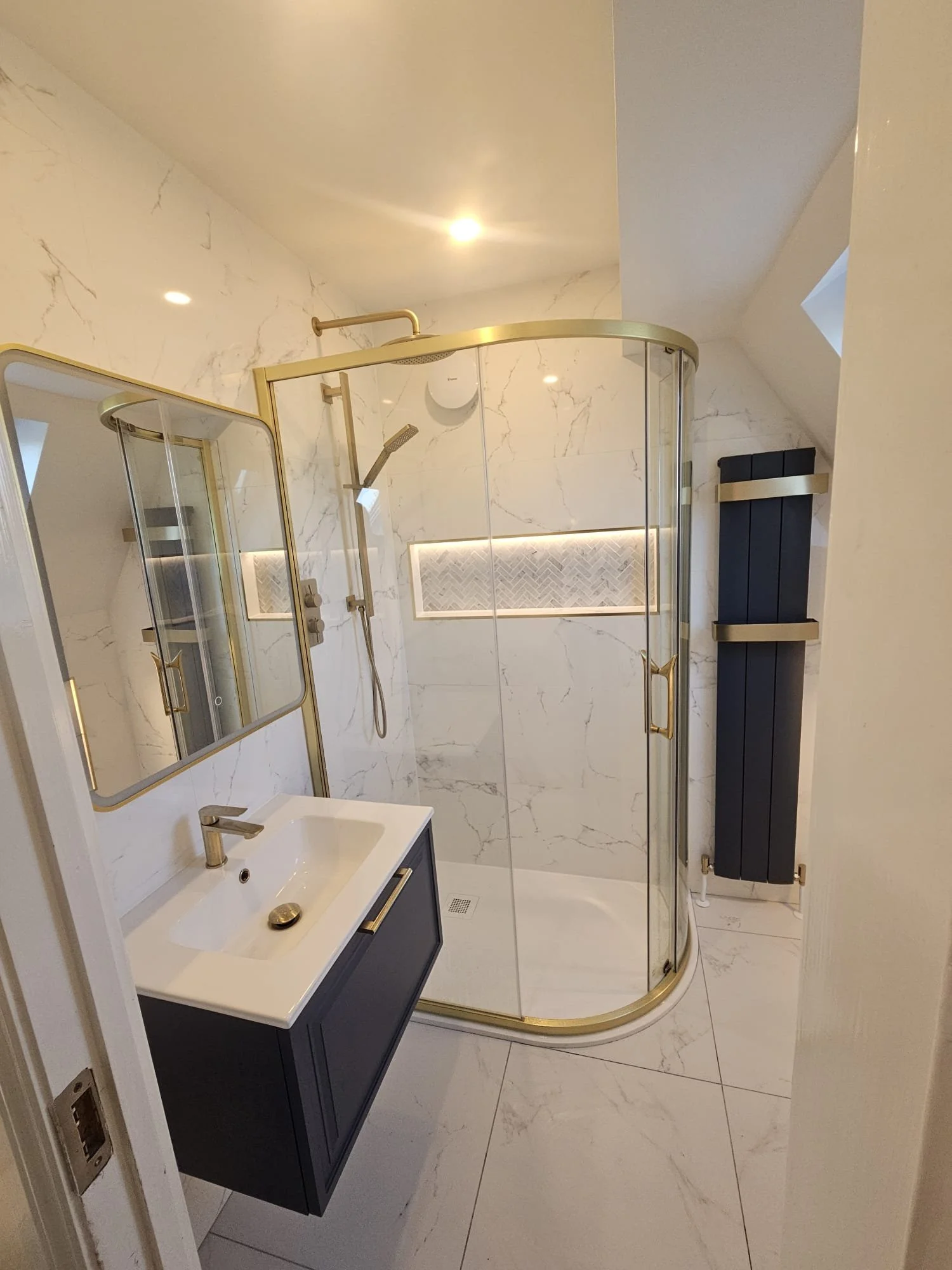 Modern bathroom with white marble walls, a curved glass shower enclosure with gold trim, a small sink with a navy blue cabinet, and a black radiator on the wall. Recessed lighting and a small window are present.