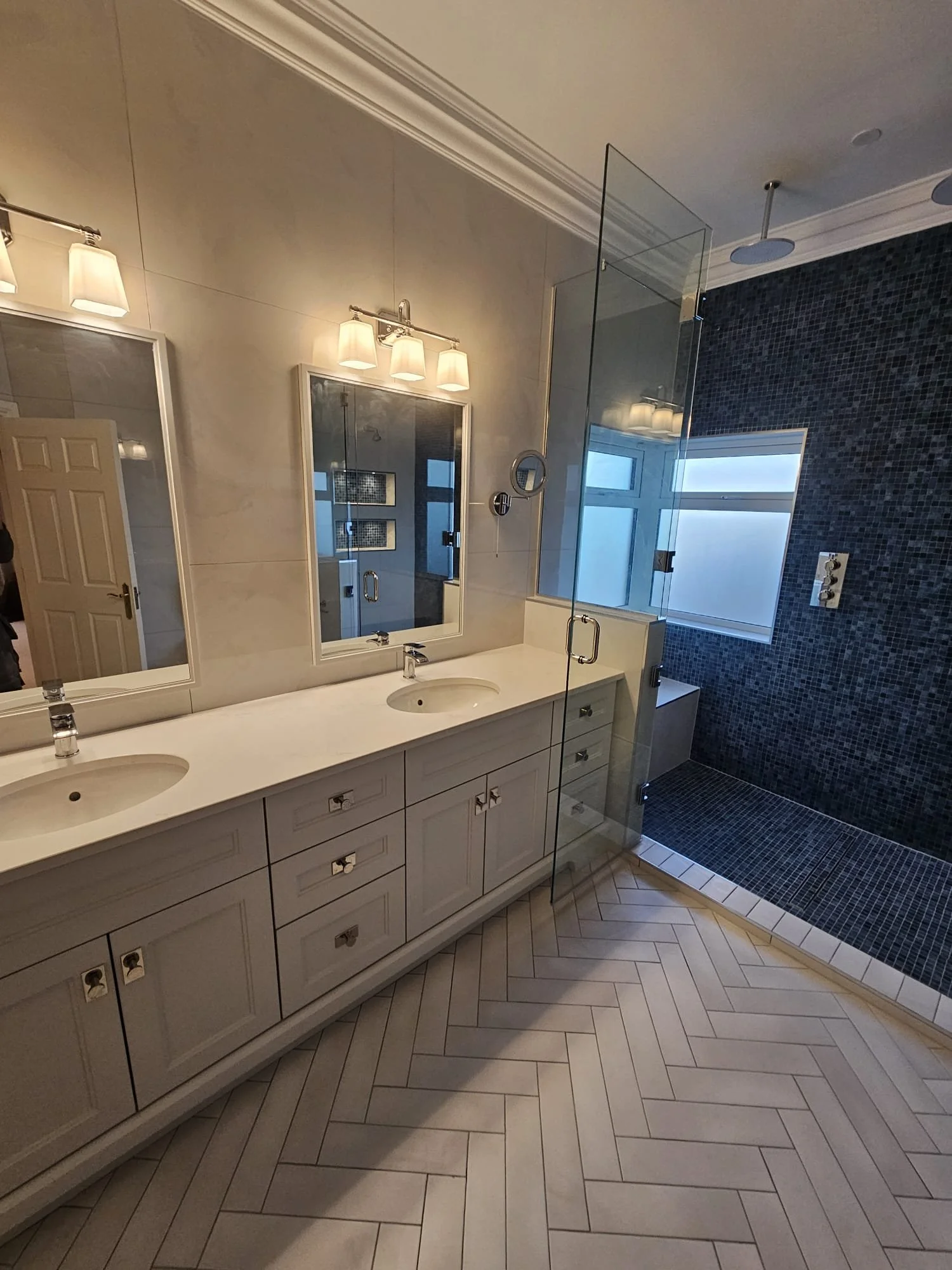 A modern bathroom with a double sink vanity, large mirror, wall-mounted lights, a glass-enclosed shower with dark tiles, a window, and beige herringbone-patterned floor tiles.