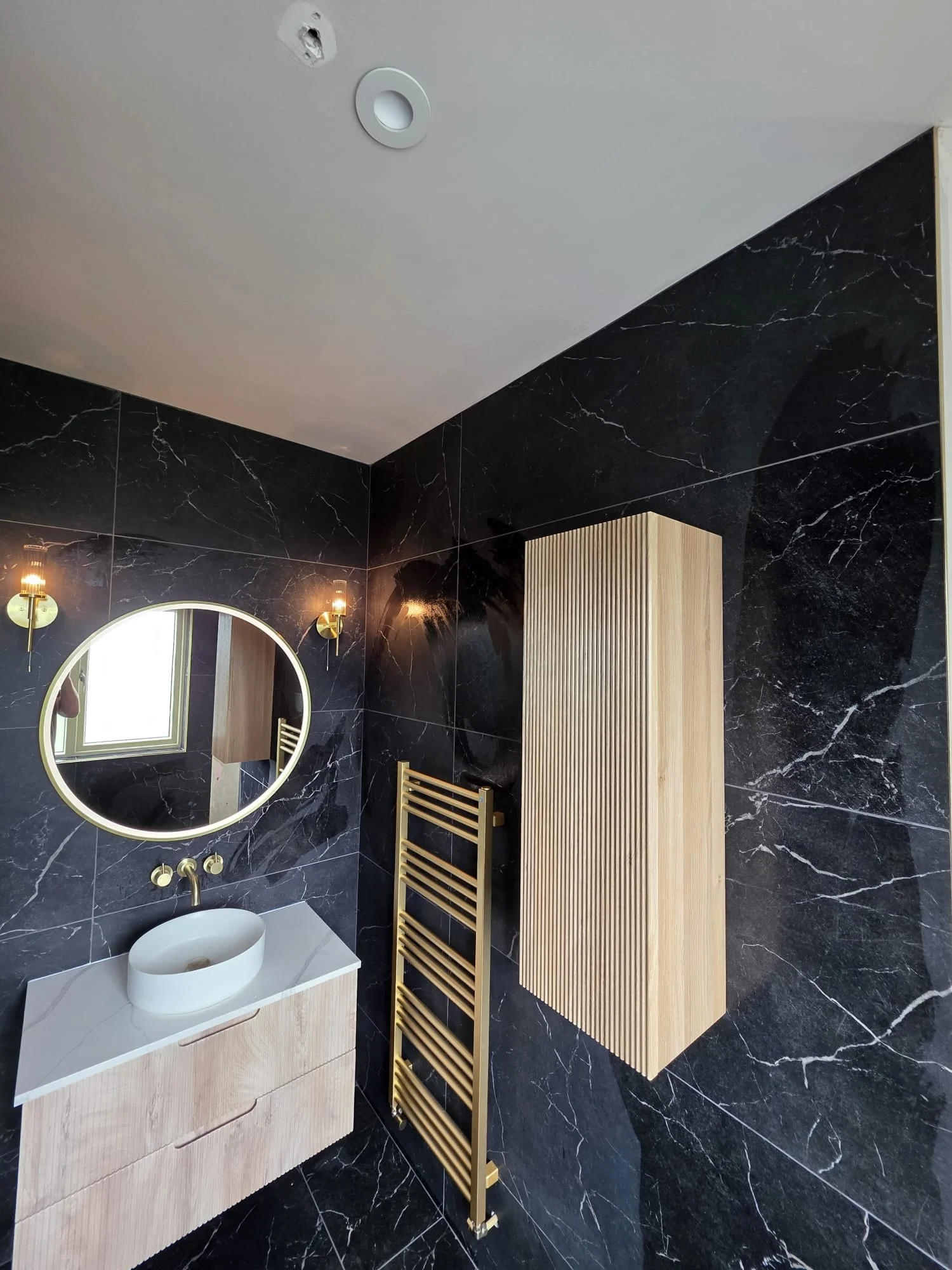 Modern bathroom with black marble walls, a white vessel sink on a wooden vanity, a round mirror, gold fixtures, a yellow wooden towel rack, and a tall light wood cabinet.