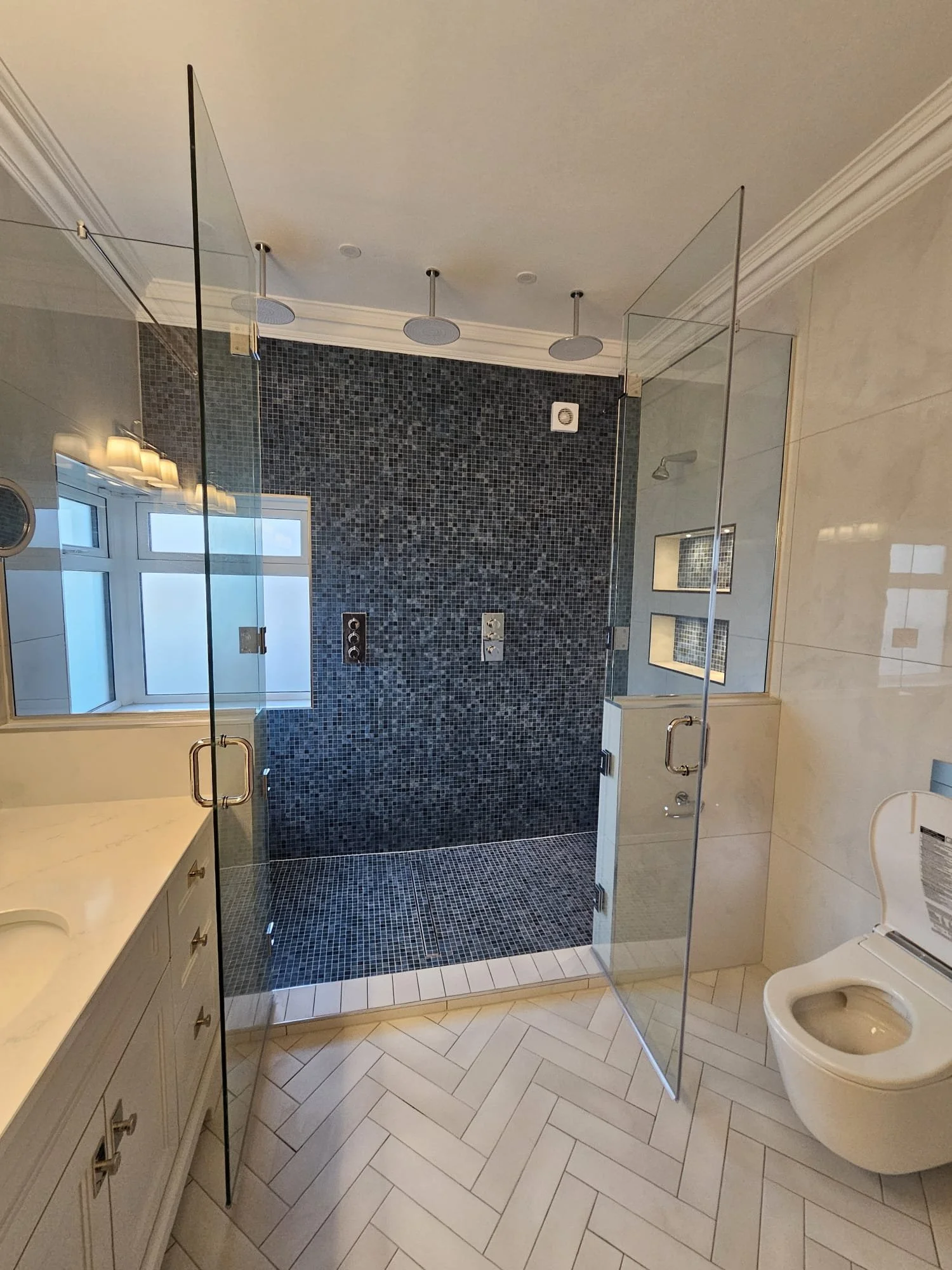 A modern bathroom with a glass-enclosed shower area that has dark blue mosaic tiles on the wall and floor. The shower features three overhead rainfall showerheads and a built-in niche with a mirror. To the left, there is a white vanity with a sink an