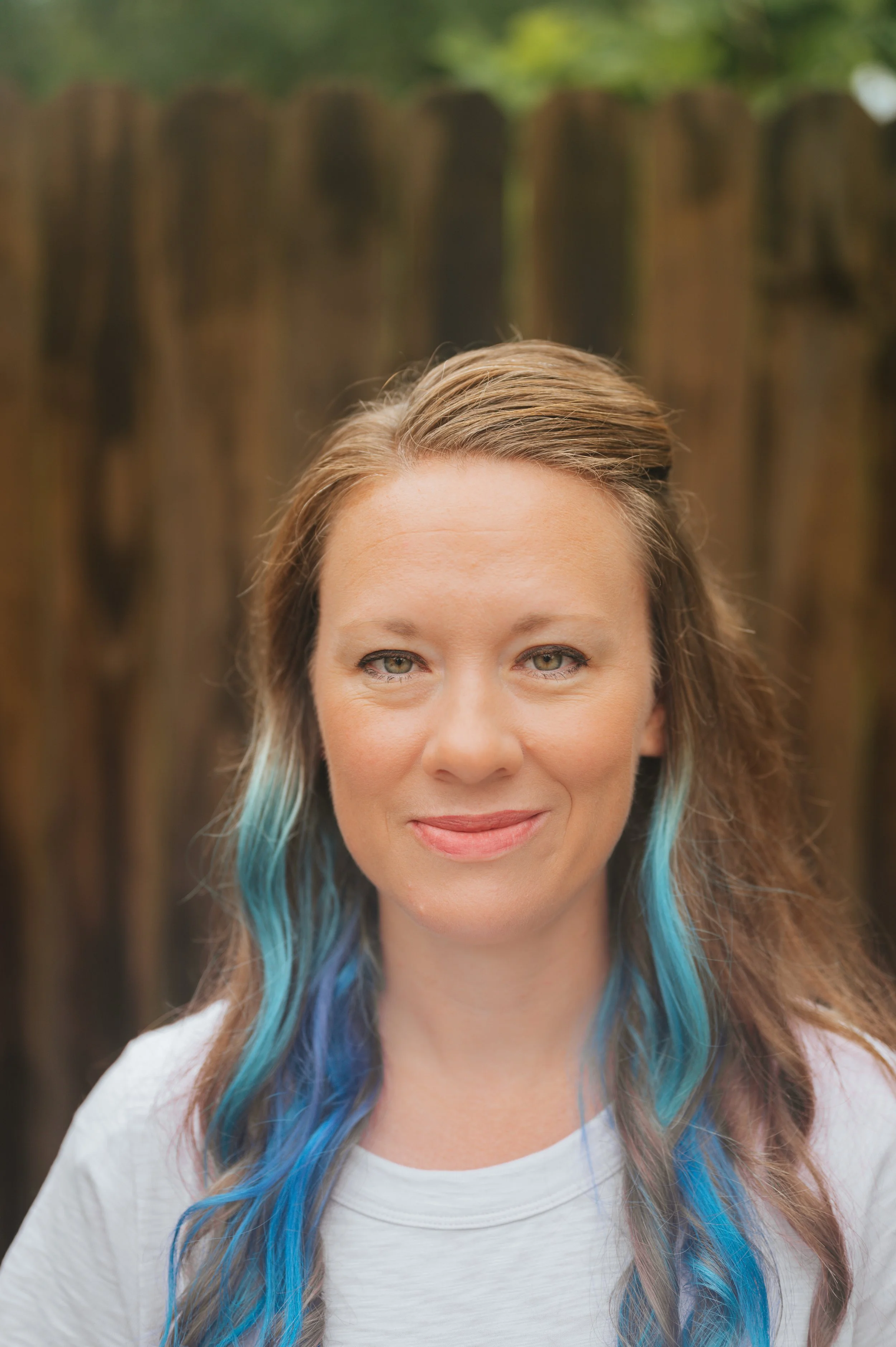Image of a woman with brown and blue hair with a scooped neck lavender shirt in front of a brown background.