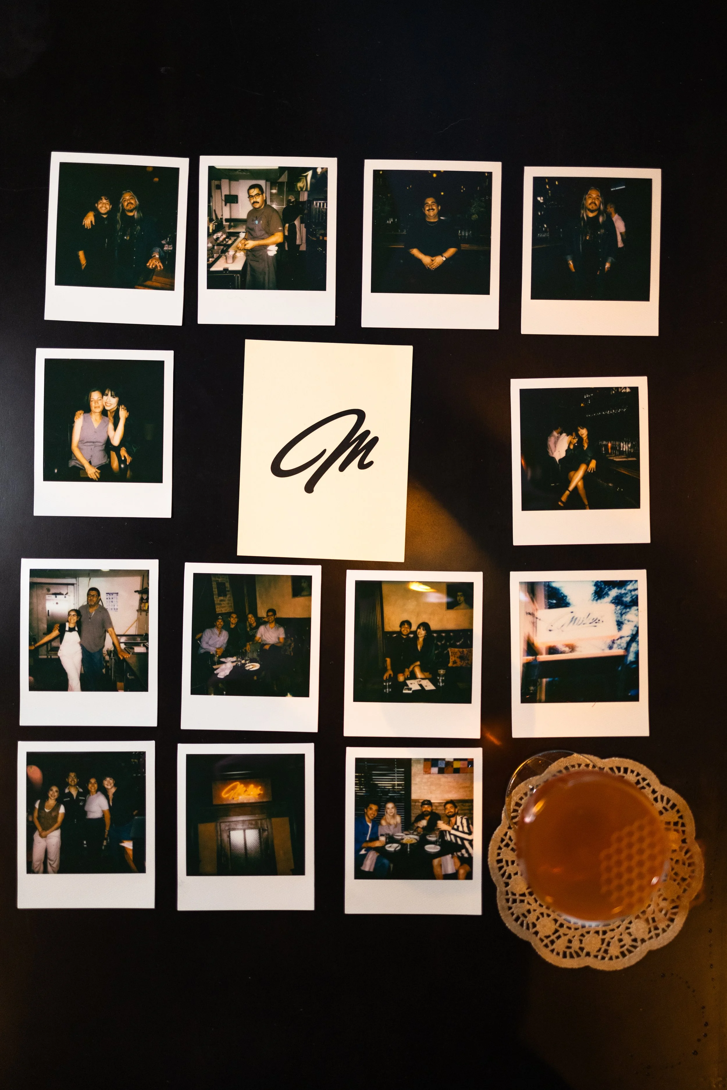 Polaroid photos displayed on a dark surface, some showing people in social settings, a sign with a signature logo, and a drink in a glass with a doily underneath.
