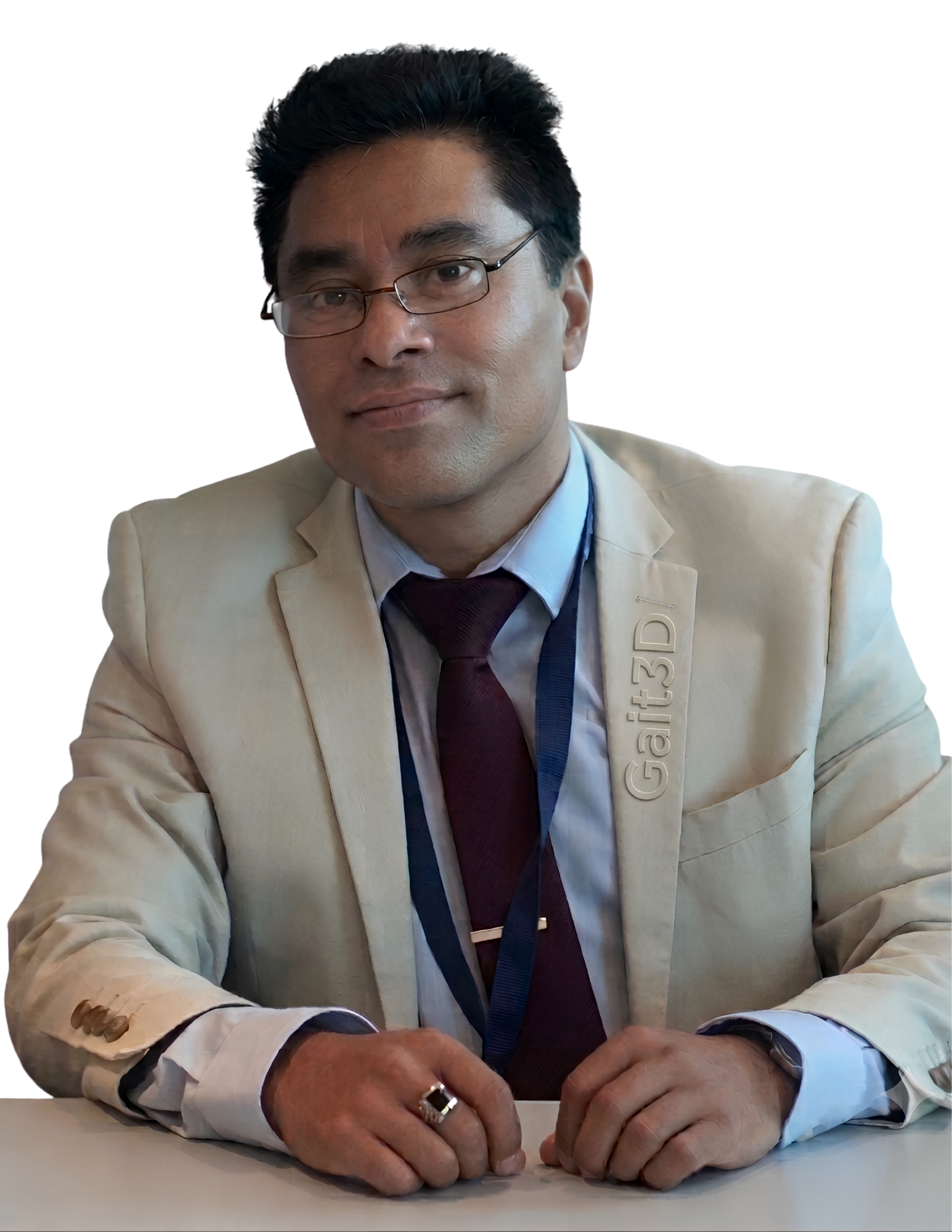 A man wearing glasses and a beige jacket sits at a table, looking into the camera with a slight smile.