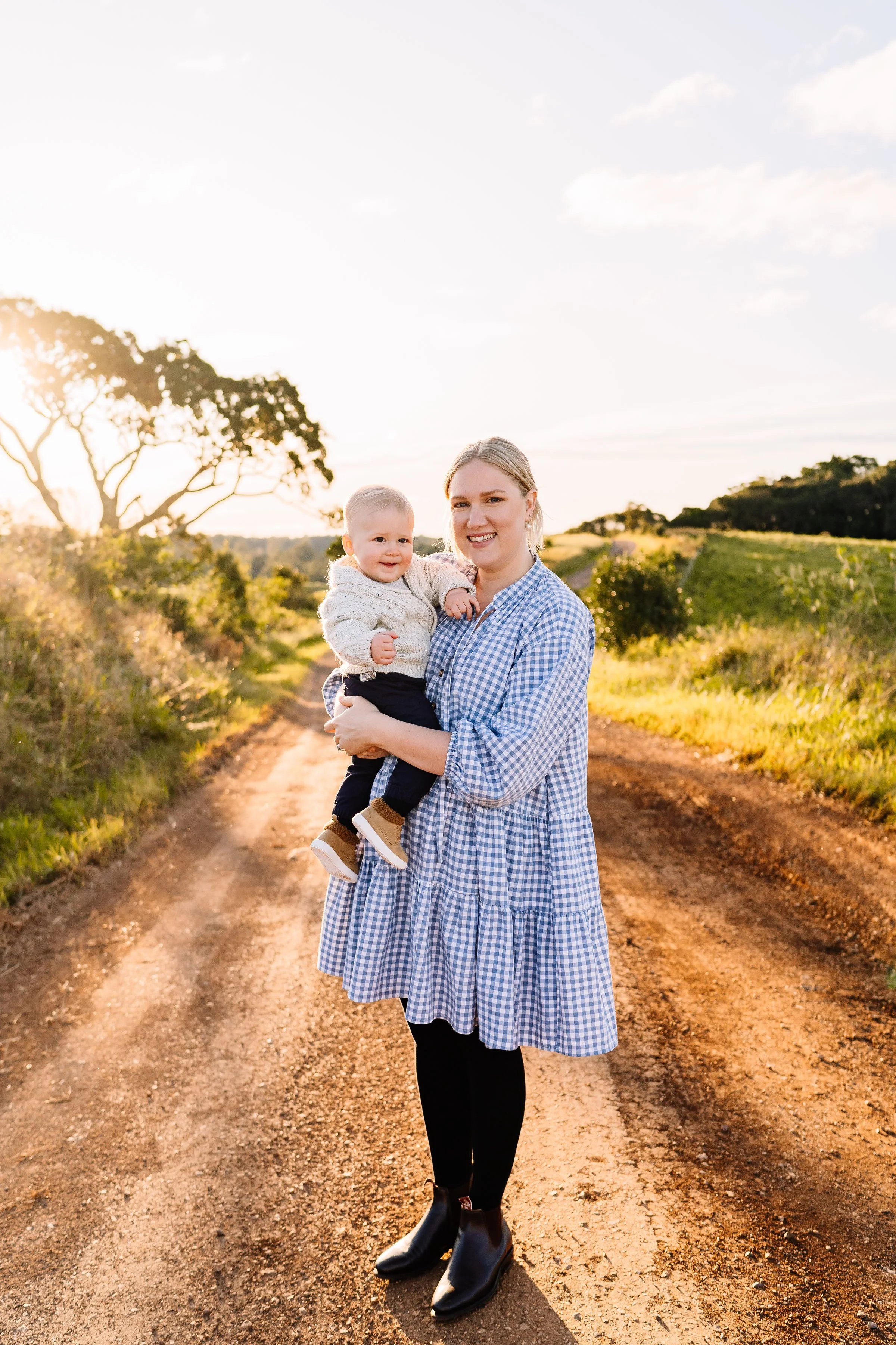 A woman holding a smiling toddler baby on a dirt path in a rural outdoor setting with trees and hills in the background during sunset.
