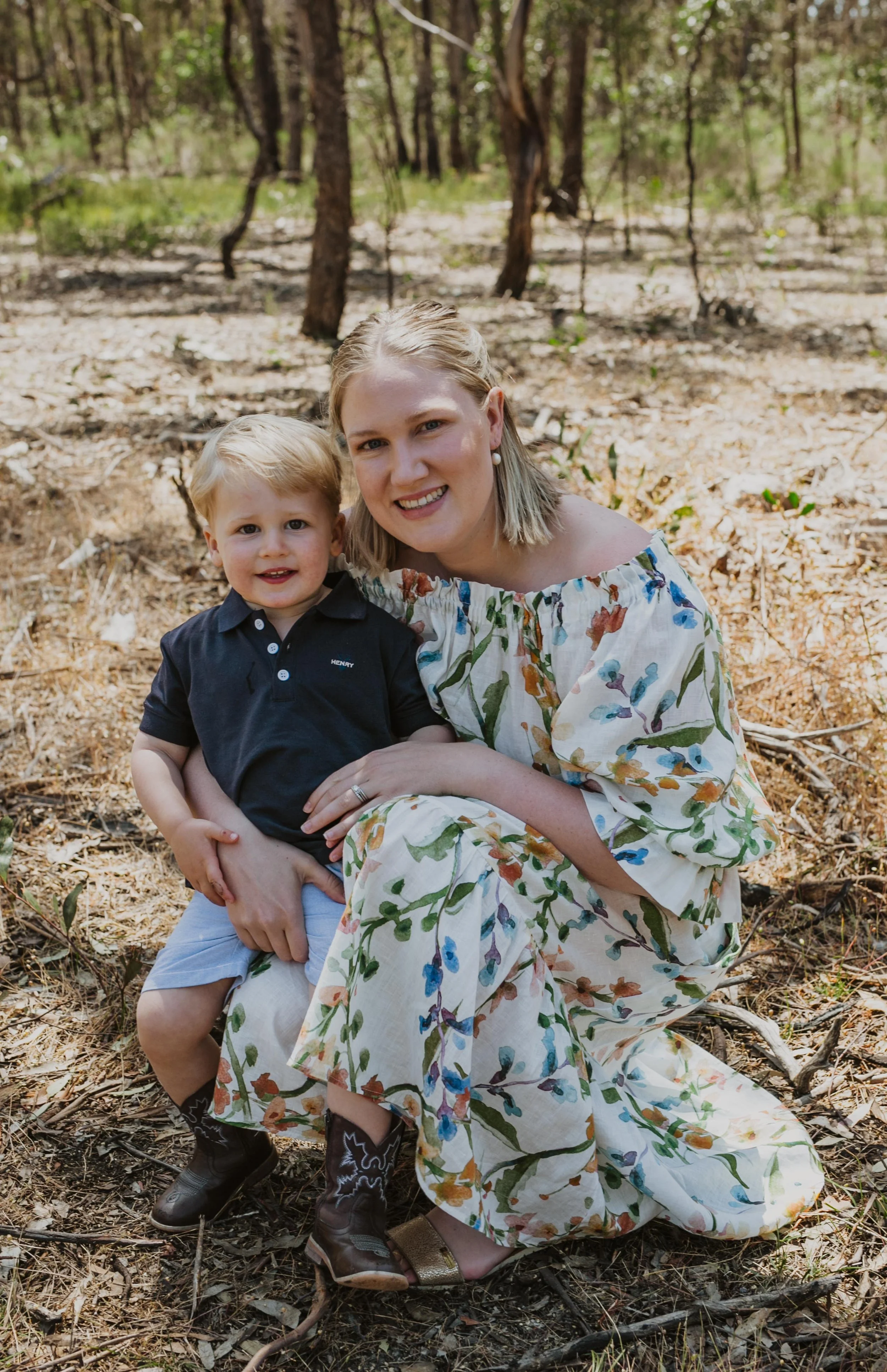 A woman and a young boy sitting on a forest floor covered with leaves and twigs, surrounded by trees, smiling at the camera.