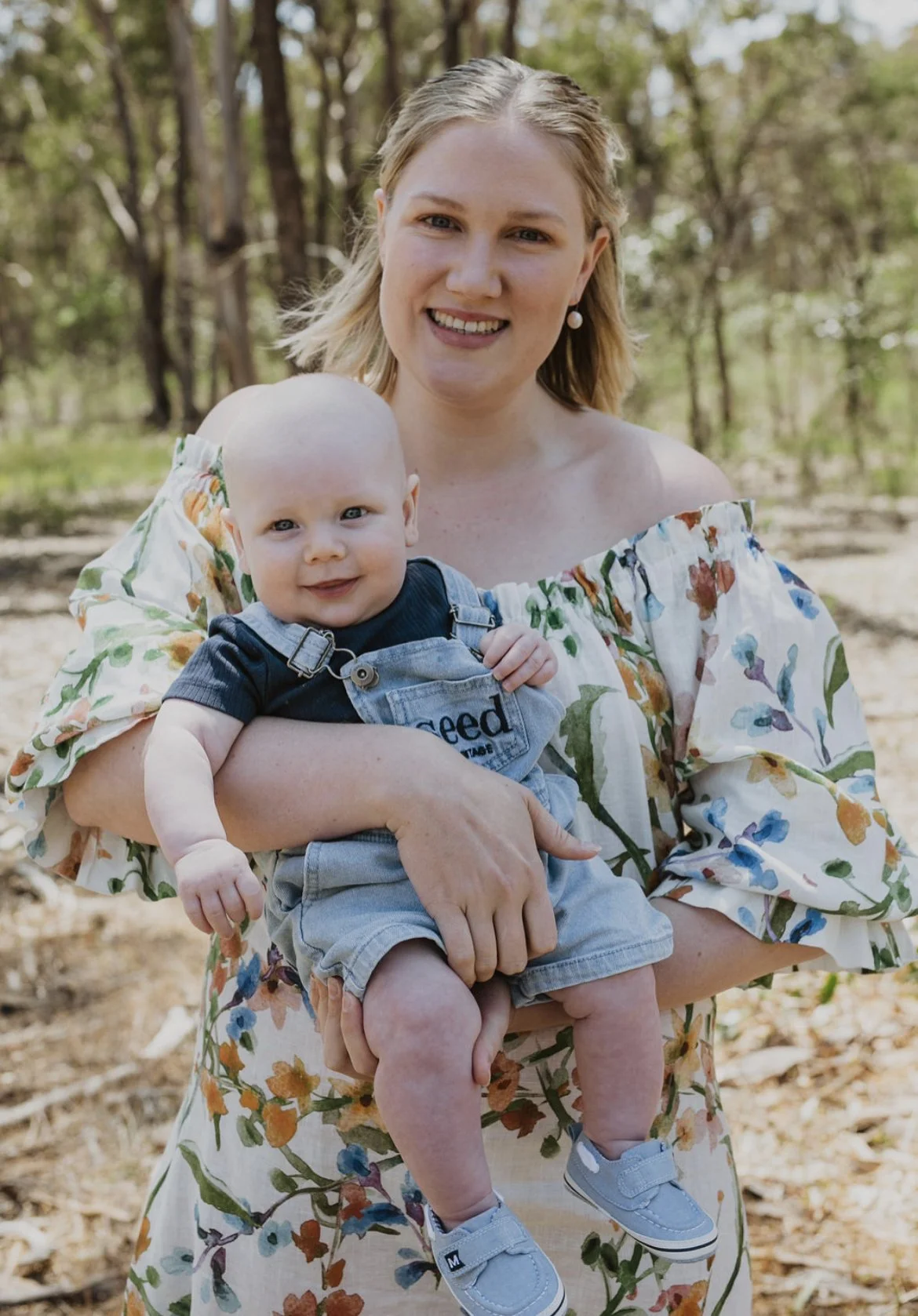 A woman holding a smiling baby outdoors in a wooded area, both dressed in casual summer clothing.