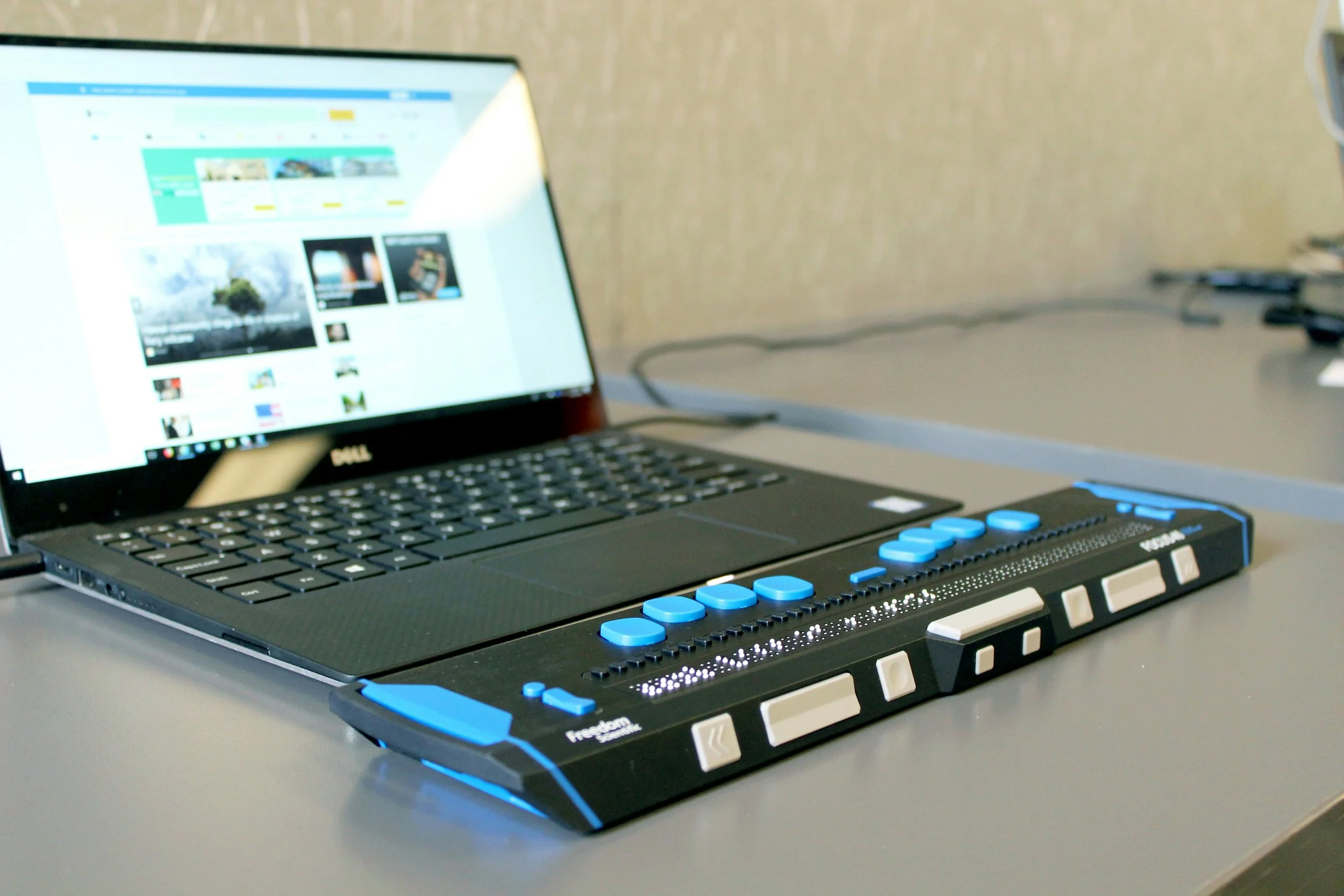 A laptop on a table displaying a webpage with images and text, connected to a control panel with blue buttons and white keys, in an office or studio setting.