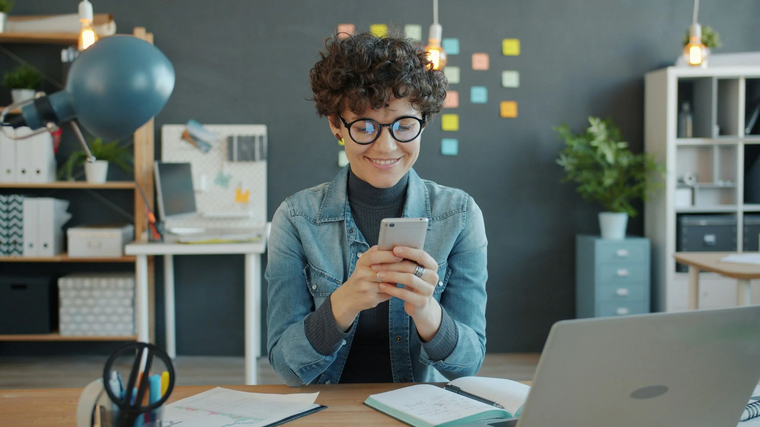 A woman with curly hair and glasses smiling while looking at her phone in a modern office with shelves, plants, and colorful sticky notes on the wall.