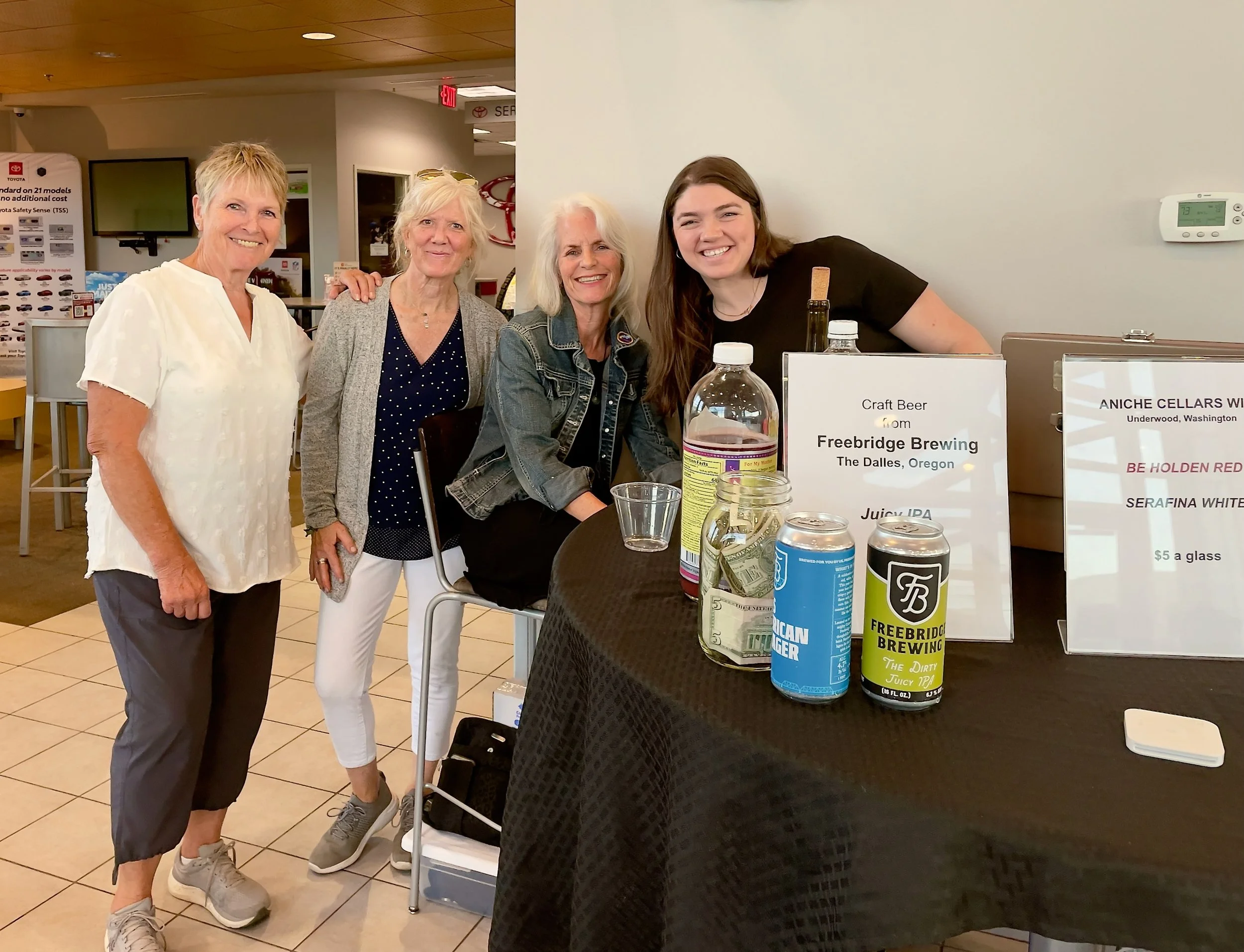 Four women standing together behind a table at an indoor event. The table displays cans of craft beer from Freebridge Brewing and Aniche Cellars. There are promotional signs on the table, and the women are smiling and facing the camera.