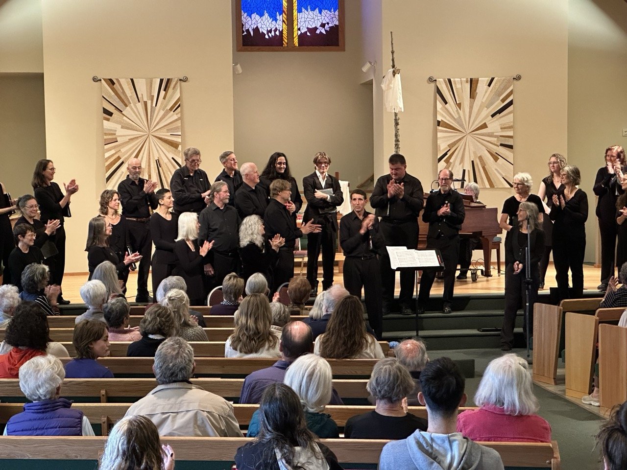 Chorus group standing on stage in a church or concert hall, clapping and smiling after a performance, with an audience seated in front of them.