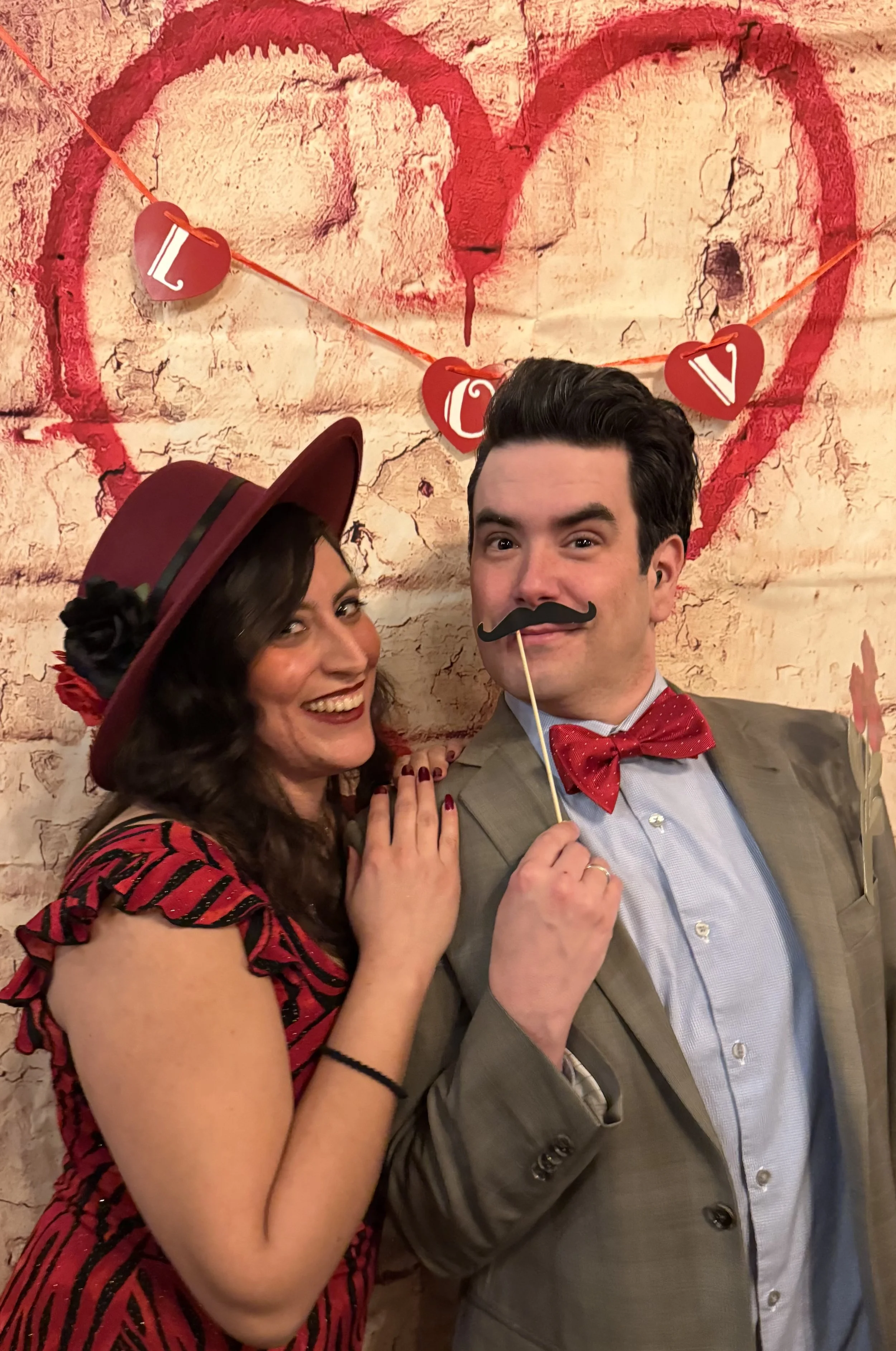 A woman wearing a red and black striped dress and a red hat with black flowers, smiling, near a man in a gray suit holding a mustache prop, both posing in front of a decorative wall with a red heart and the word 'LOVE' on a banner.