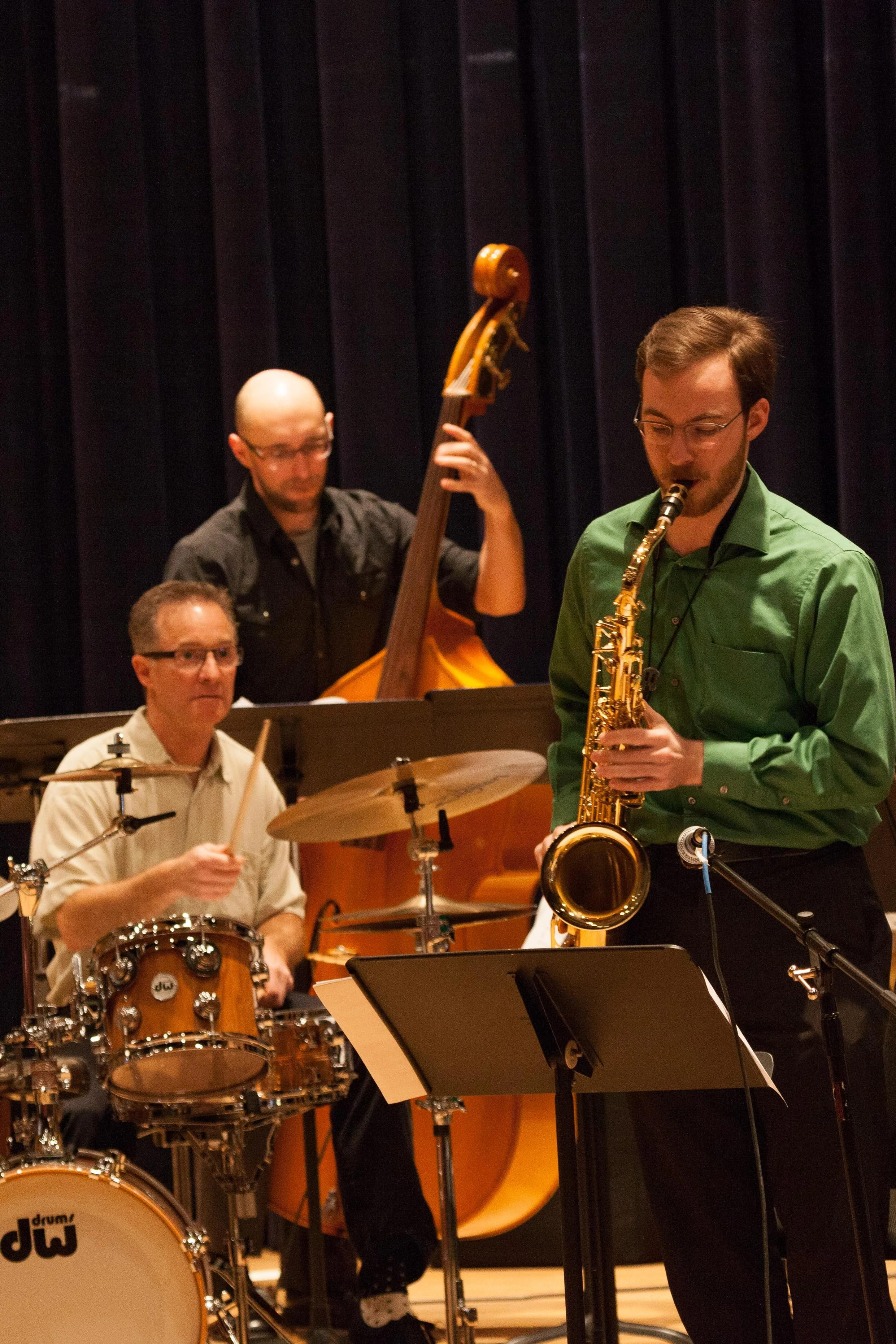 A jazz band performing on stage, featuring a drummer, a bass player, and a saxophonist. The drummer is in the foreground, the bassist is slightly behind, and the saxophonist is to the right, all focused on their instruments.