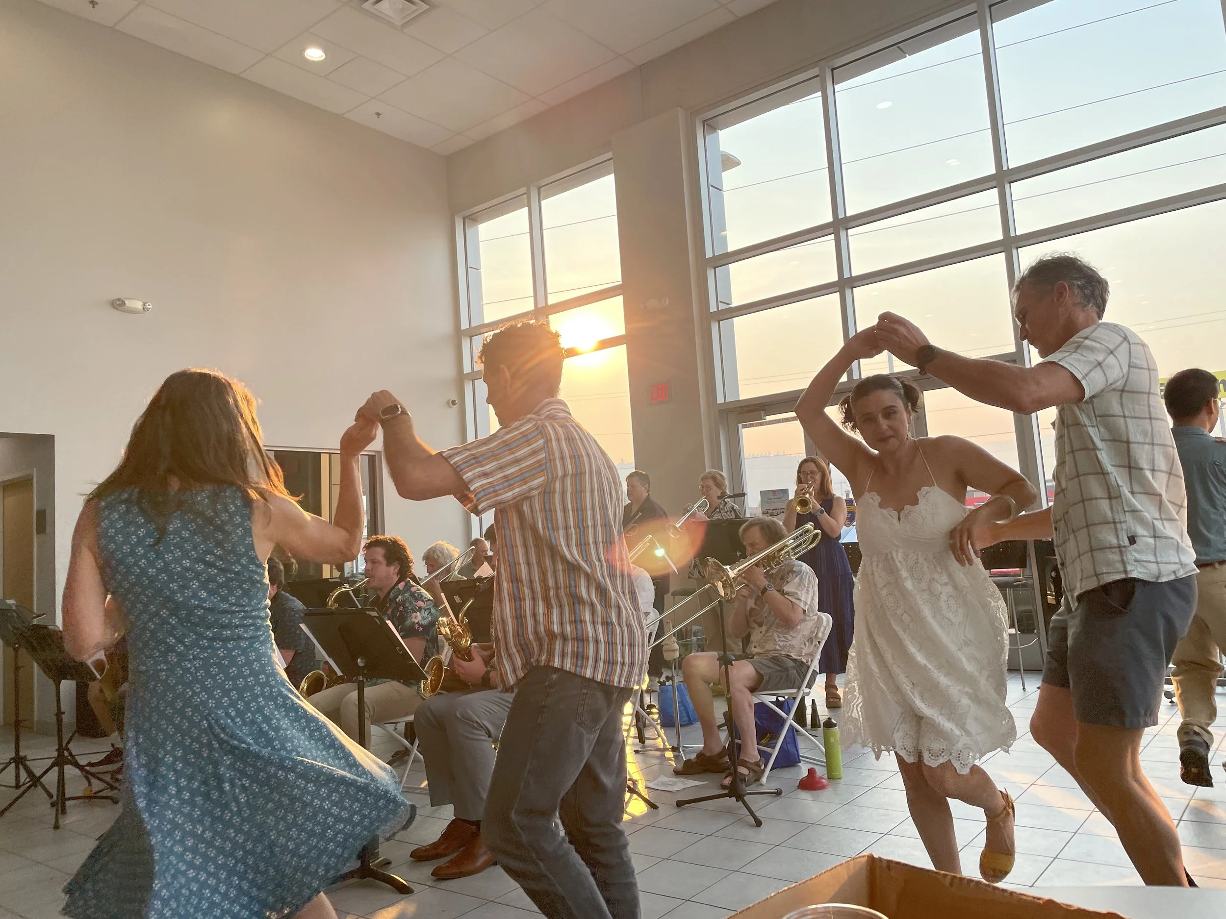 People dancing and playing musical instruments in a brightly lit room with large windows during sunset.