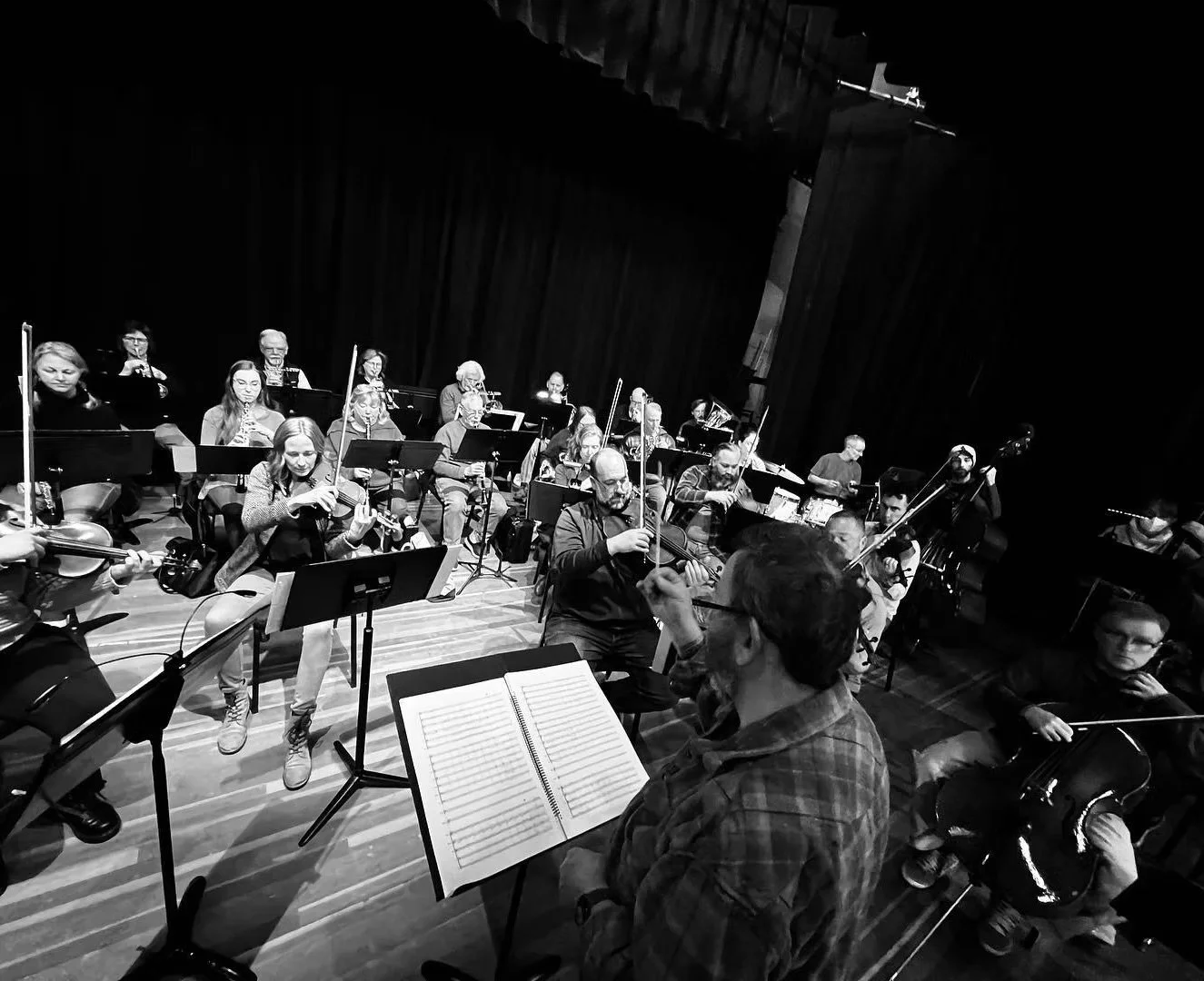 A black and white photo of an orchestra practicing in a theater, with musicians playing violins, cellos, drums, and other instruments, led by a conductor.