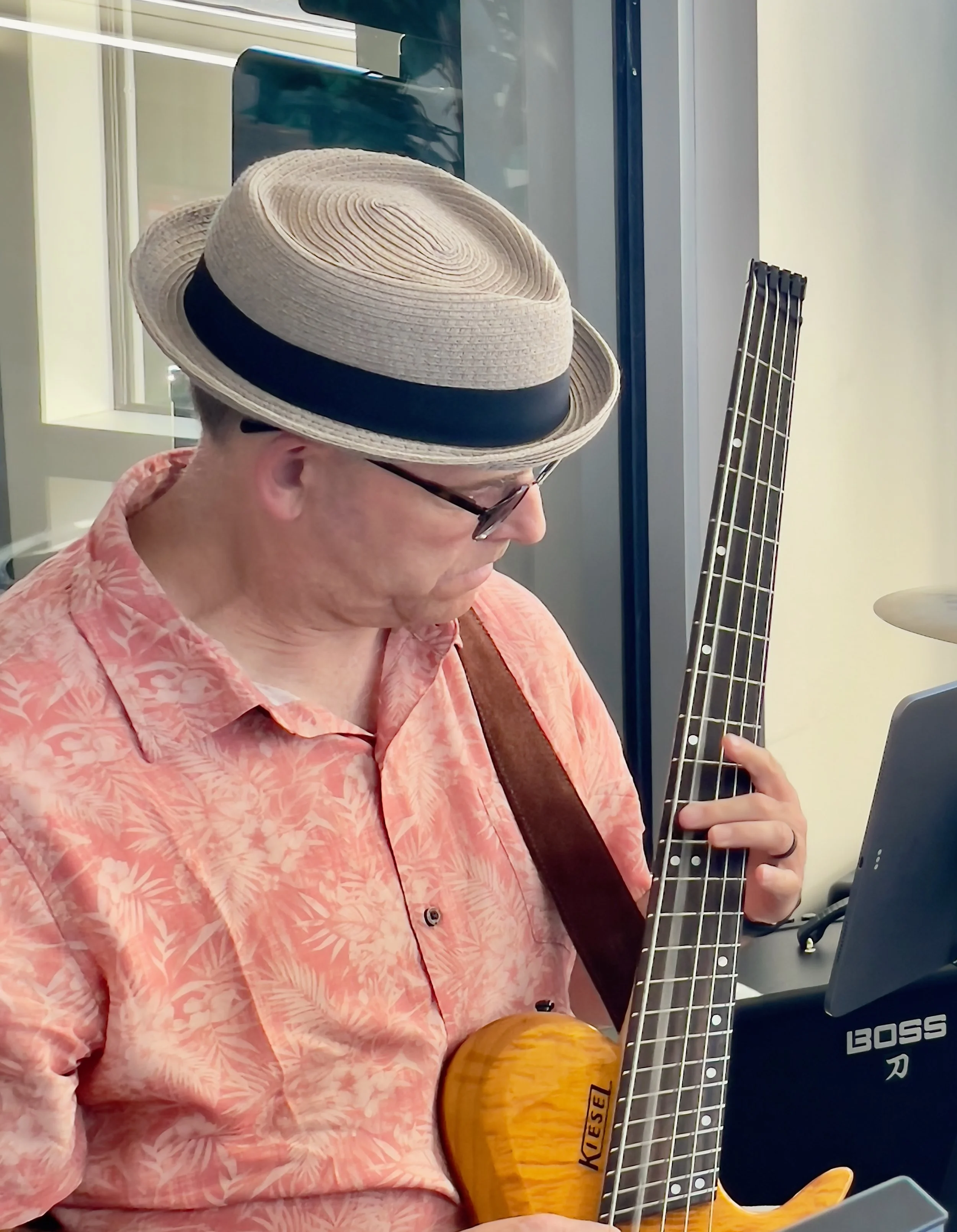A person with light skin wearing a straw hat with a black band, glasses, a pink floral shirt, and holding a yellow bass guitar with a wood grain finish, sitting indoors near a window.