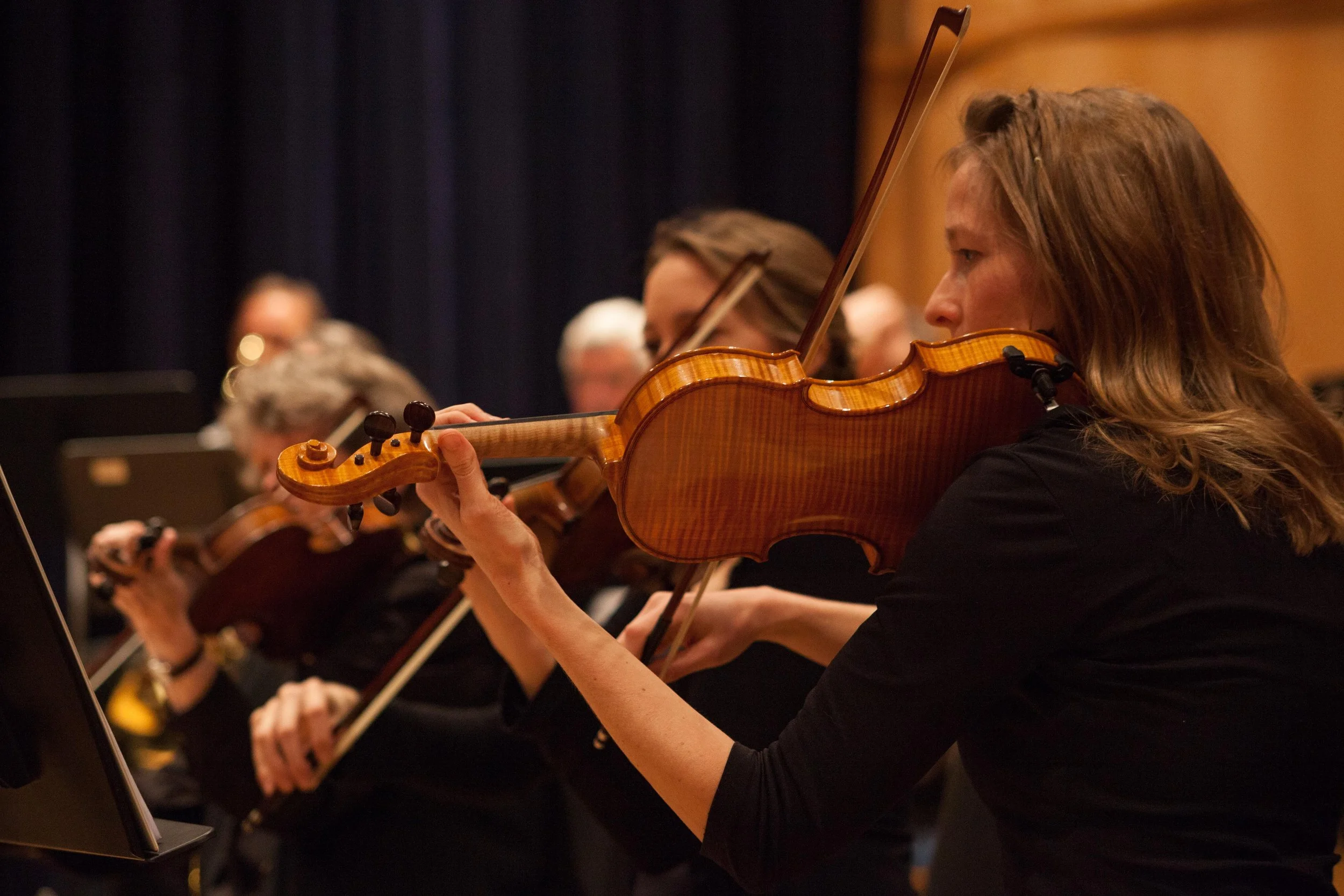 Women playing violins in an orchestra, seated in a row, with music stands and black curtains in the background.