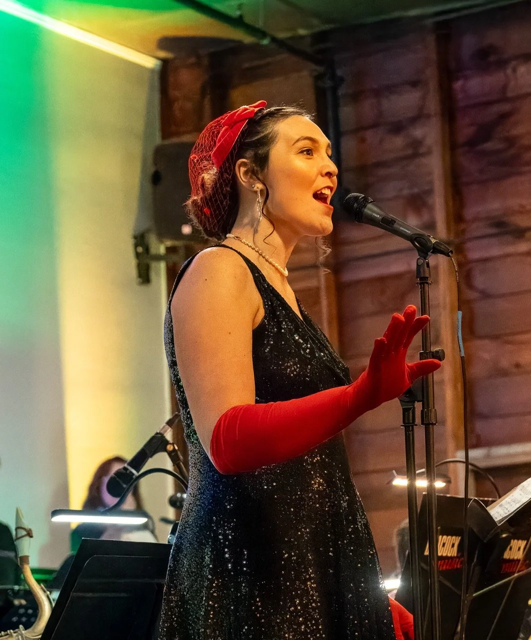 Woman singing at microphone wearing a black sequined dress, red gloves and headscarf, in a cozy wooden venue.