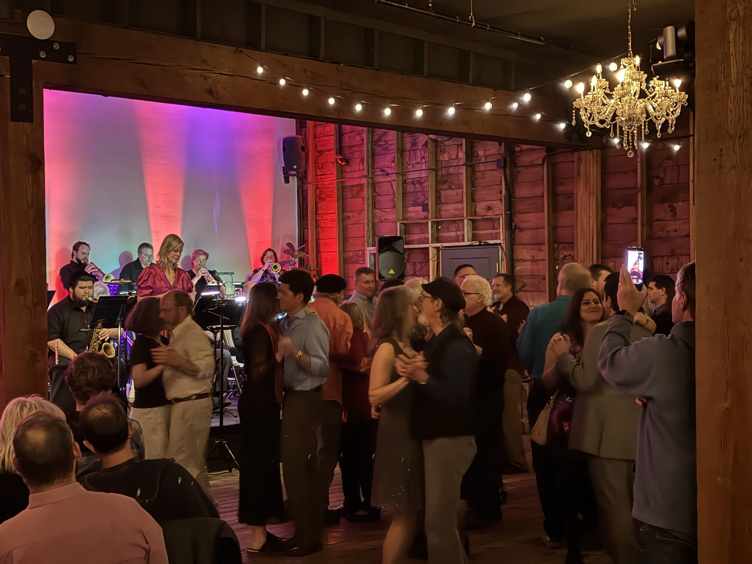 A lively indoor event with a jazz band performing on stage, surrounded by a crowd of people socializing and dancing. The venue has wooden walls, string lights, and a chandelier, creating a warm, festive atmosphere.