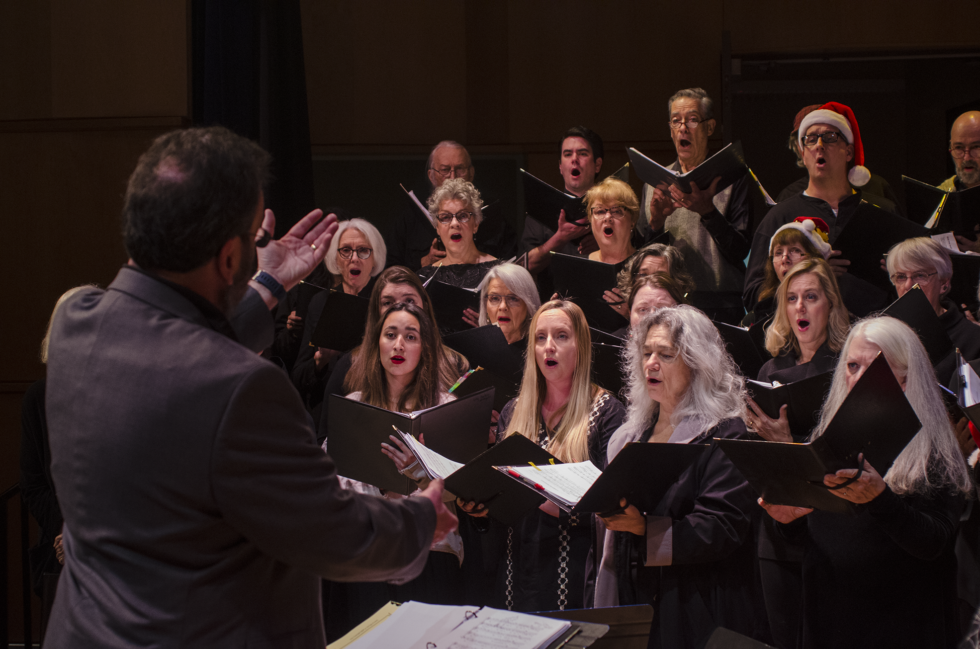 A choir of men and women singing, led by a conductor. Some choir members are wearing Santa hats, and the conductor has his hand raised.