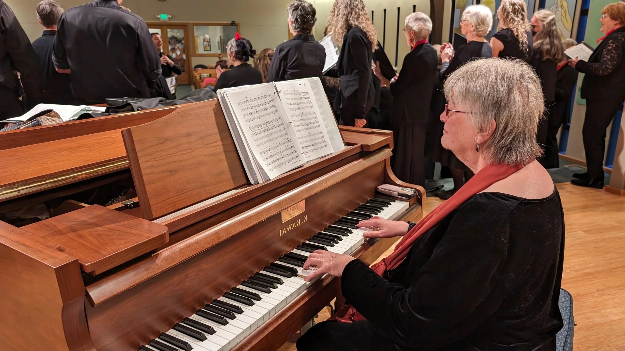 A woman playing a wooden upright piano in a room filled with people, some standing and some sitting, singing or listening, during a musical or religious gathering.