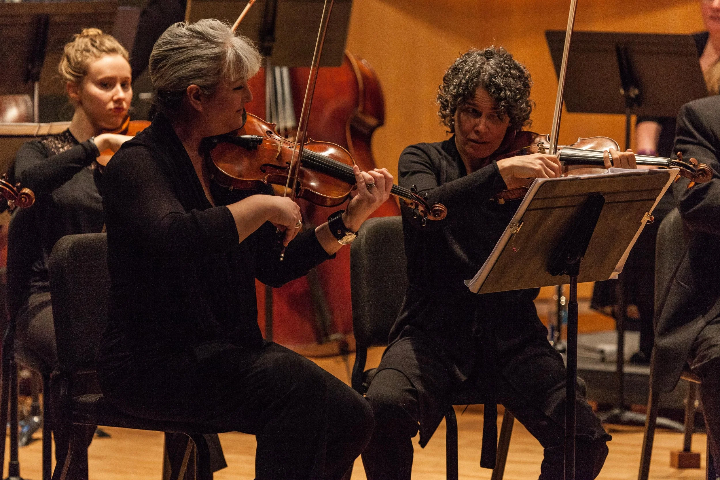 Three women playing violins in an orchestra. The woman on the left has gray hair, and the woman on the right has curly hair. They are seated, reading music, and focusing on their instruments. A young woman with blonde hair is visible in the background, also playing the violin.