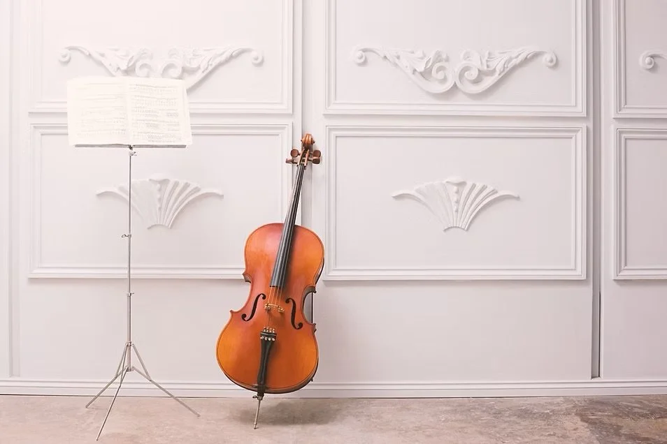A classical cello leaning against an ornate white wall, with a music stand holding sheet music nearby.