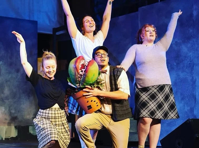 Four performers on stage, one holding a large puppet with a plant pot, and three raising their arms, smiling or singing.