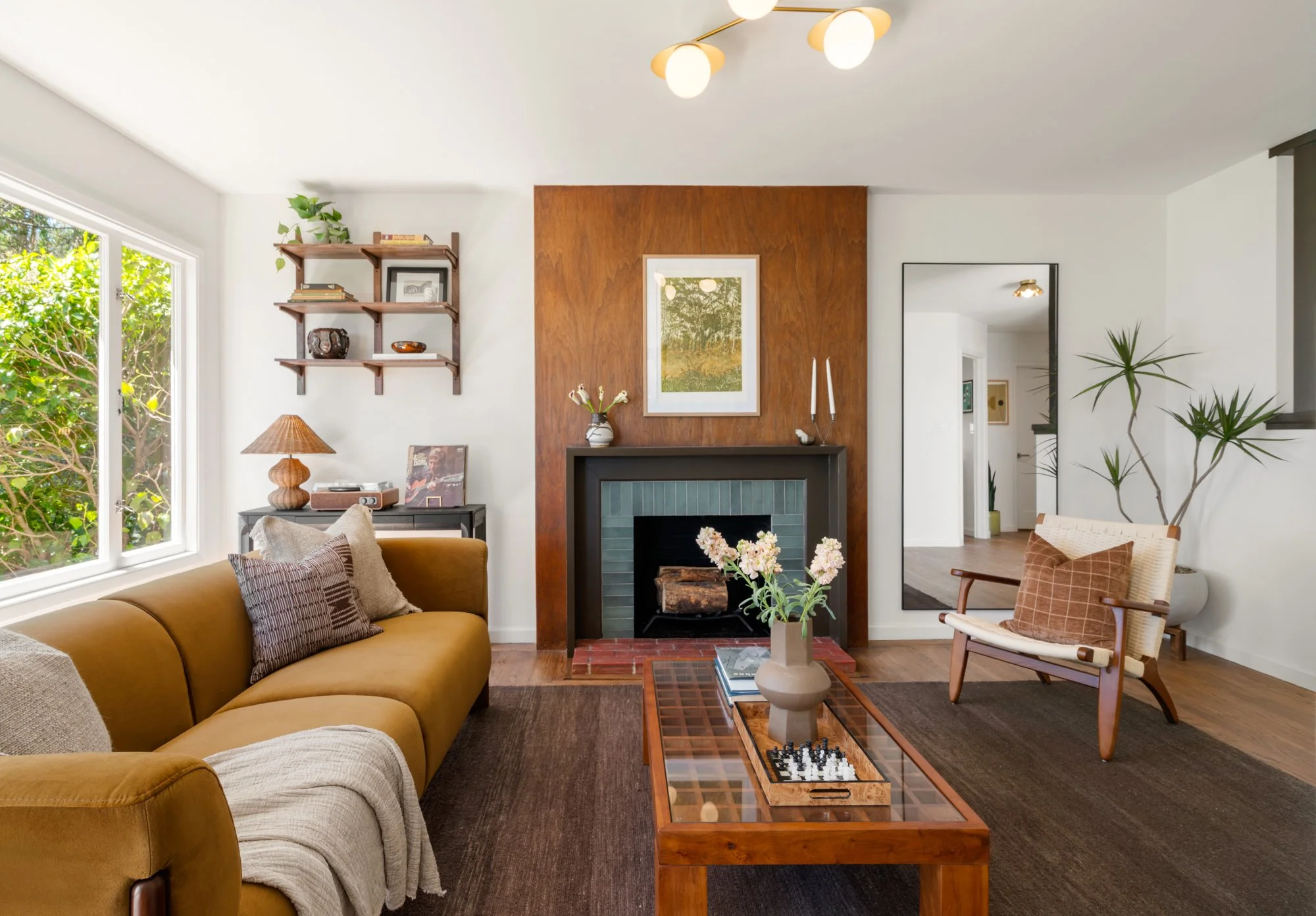 Living room with a mustard-colored sofa, patterned cushions, a wooden coffee table with a chess game, a fireplace with a black frame, a large mirror, a wooden wall with a framed picture, a vase with flowers, a shelf with books and decor, and a window showing trees outside.