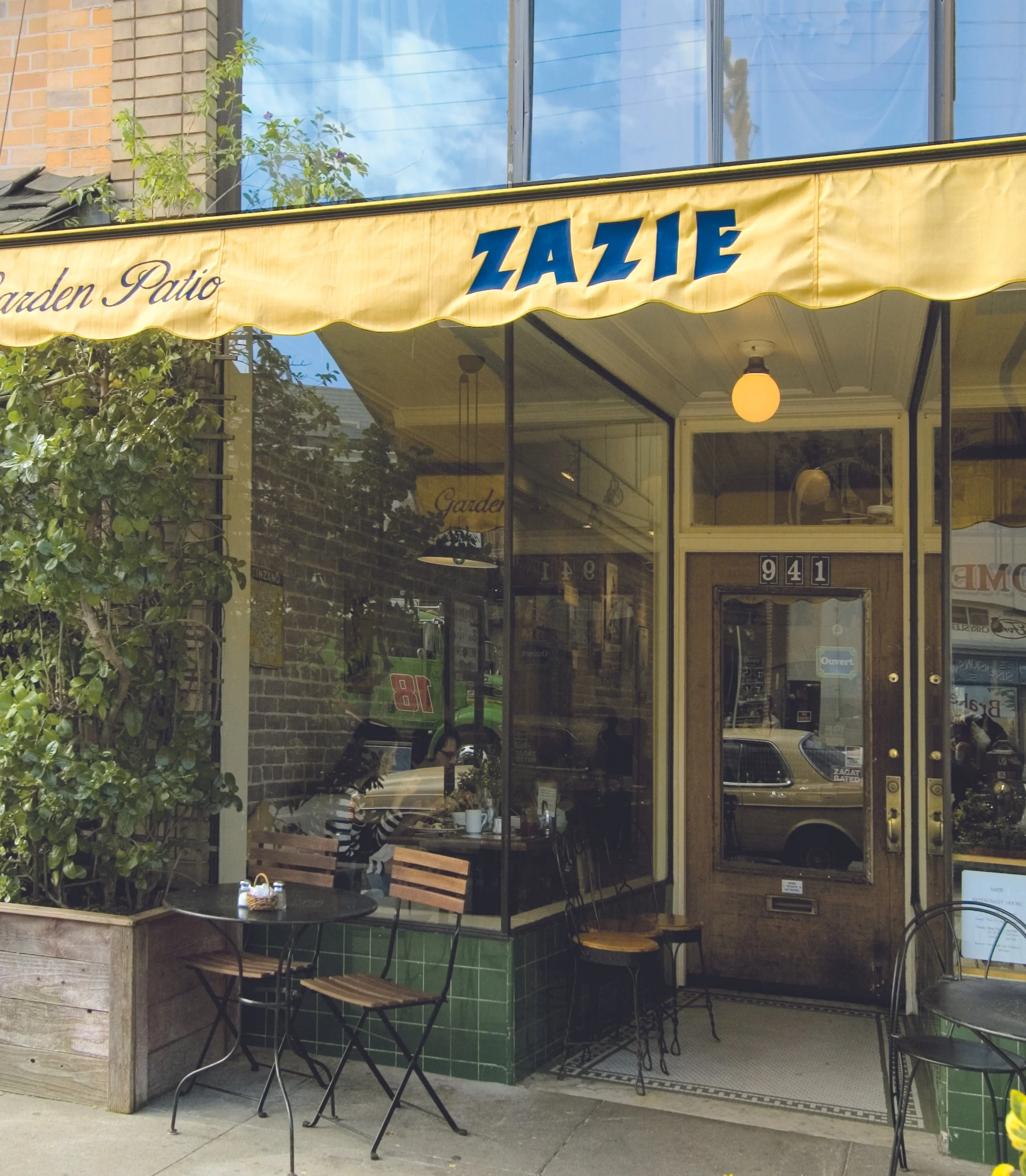Restaurant storefront with a yellow awning displaying the name 'ZAZIE' and the words 'Garden Patio'. Large glass windows reveal a cozy interior with tables, chairs, and a person sitting inside. Outdoors feature small round tables and chairs, some wit