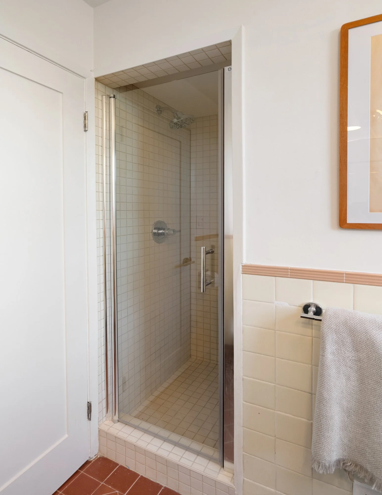 A corner shower with glass door and beige tiled walls inside a bathroom, next to a white door, with a towel hanging on a black towel ring on the tiled wall.