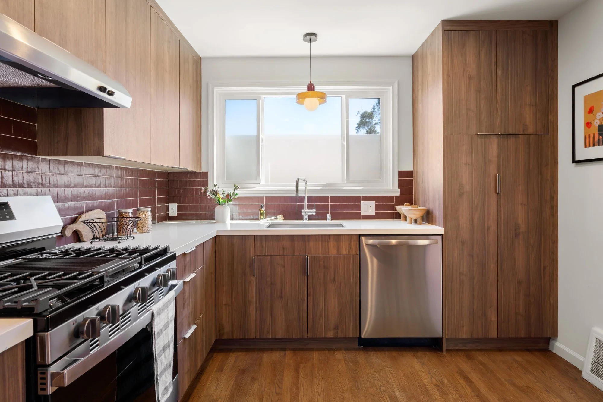 A modern kitchen with wooden cabinets, a white countertop, a gas stove, a sink with a window above, and a pendant light hanging from the ceiling.