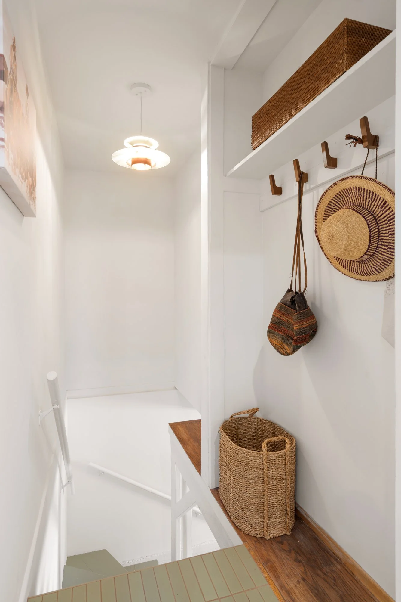 A small entryway with white walls and a hanging light fixture. There are wooden and woven baskets, a striped hat, and a bag hanging on hooks. A white shelf with a woven box is above the hooks. The floor is a mix of white tiles and dark wood.
