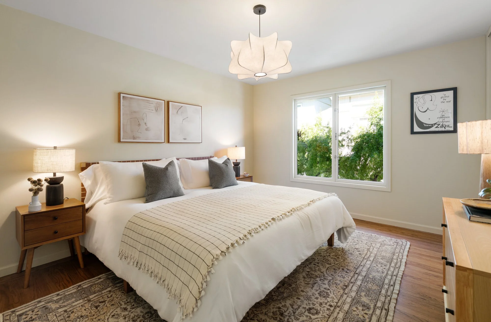 A cozy bedroom with a neatly made bed, white linens, and gray pillows. A wooden nightstand with a black table lamp and decorative items sits beside the bed. There are two framed abstract art pieces on the wall above the bed, and a window showing gree