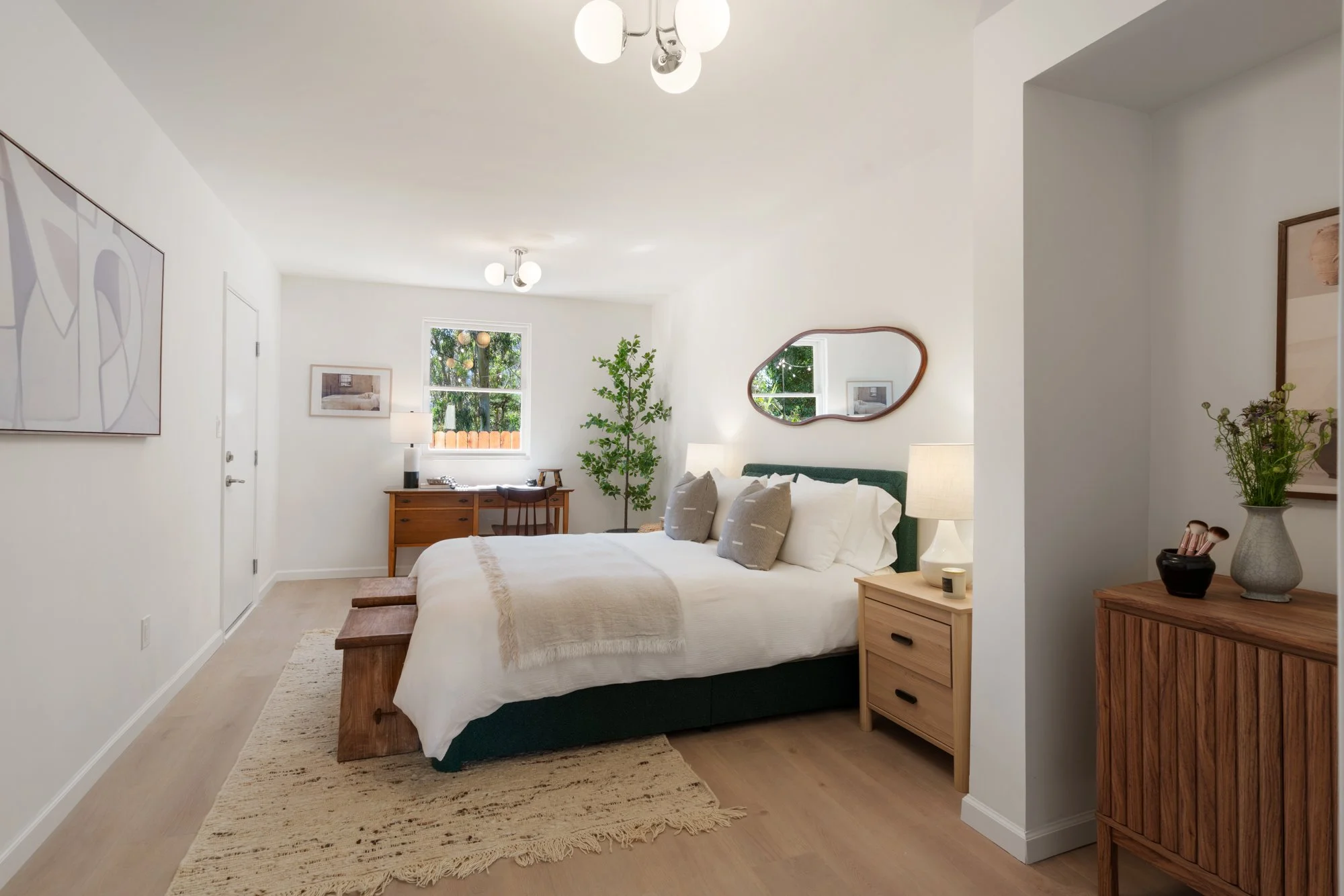 A cozy and modern bedroom with a large bed, white bedding, and decorative pillows. There are two wooden nightstands with lamps, a wooden desk with a chair, a mirror on the wall, a framed picture, a potted plant, and ceiling lights. The room has a window with a view of greenery outside.