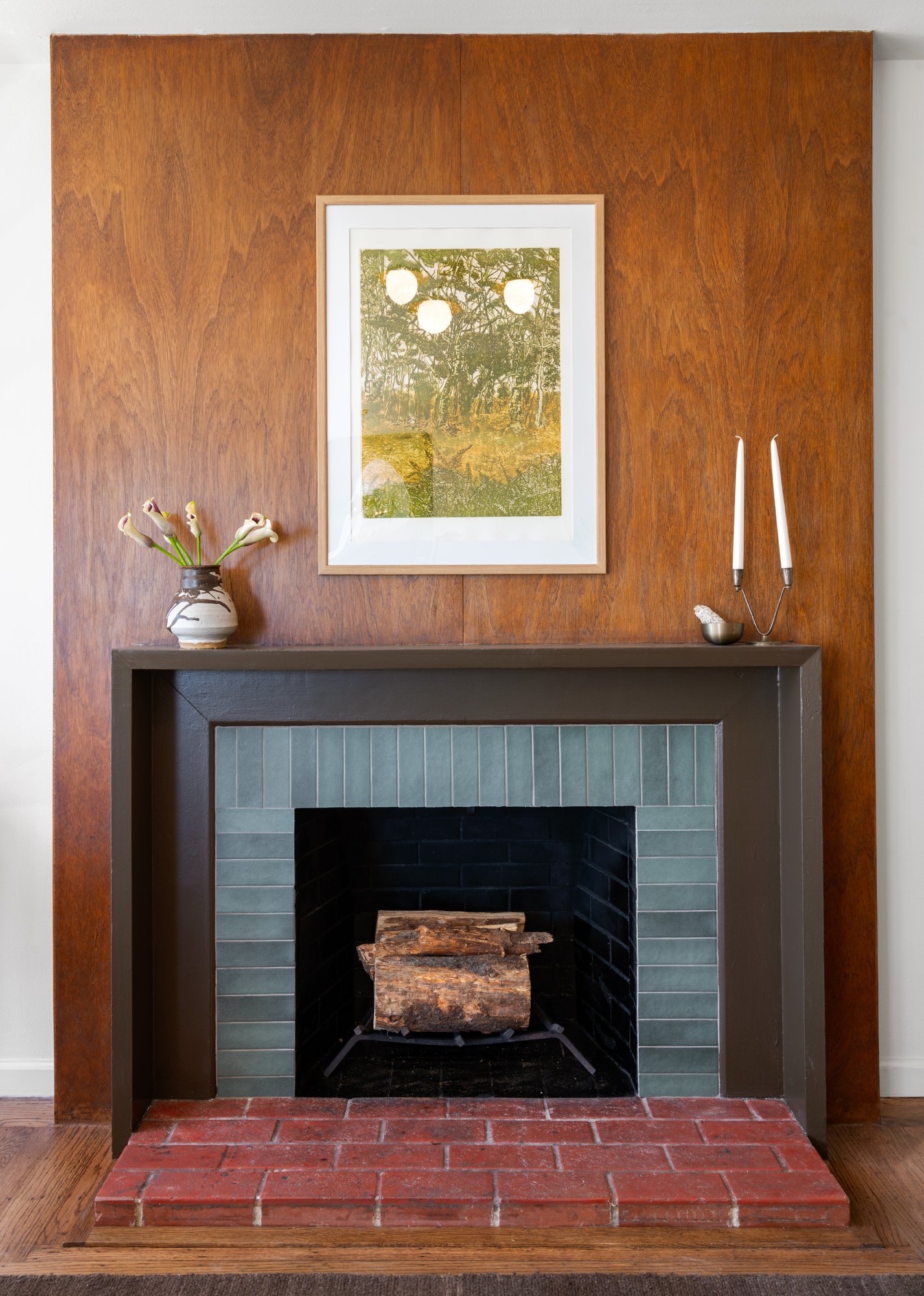 A decorative fireplace with a wood mantel, gray tile surround, and red brick hearth. A small vase with calla lilies and a candle holder with white candles are on the mantel. A framed picture of a forest scene hangs above.