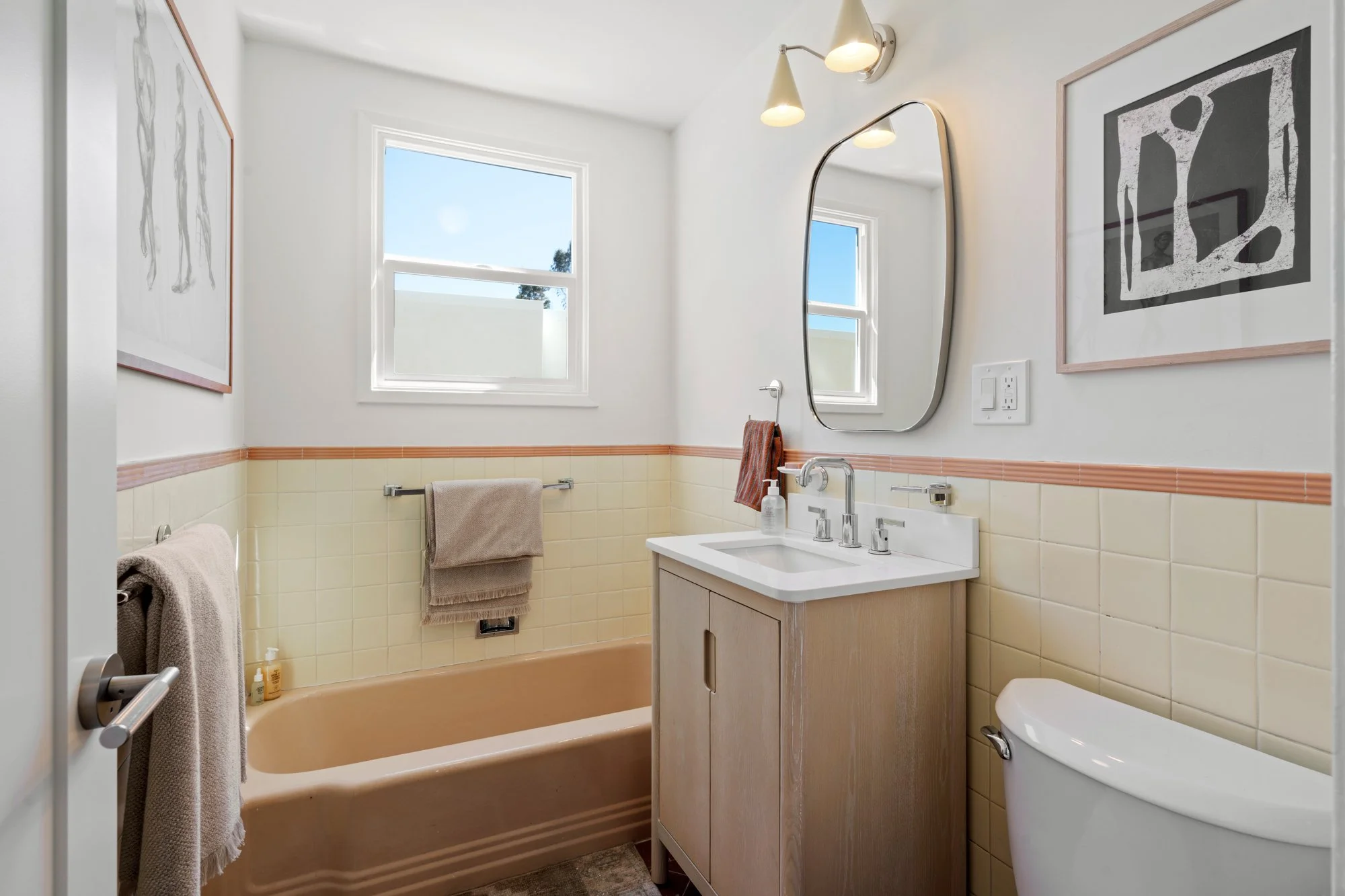A bathroom with a beige bathtub, a small vanity with a sink, a mirror, and two framed art pieces on the white walls. There are two windows, one above the bathtub and one above the sink, and beige and light brown towels.