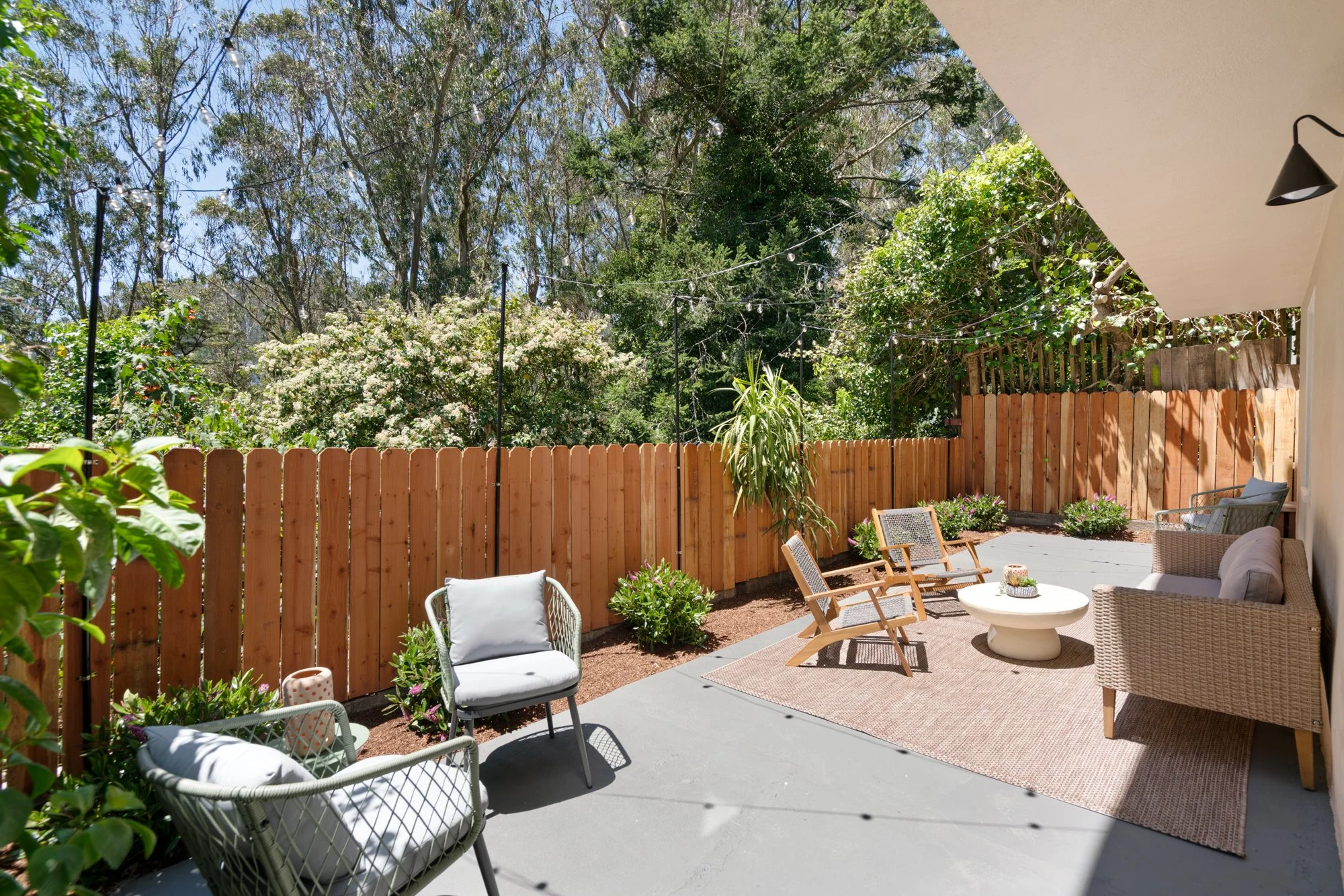 Backyard patio with outdoor furniture, green plants, a wooden fence, string lights, and trees in the background.