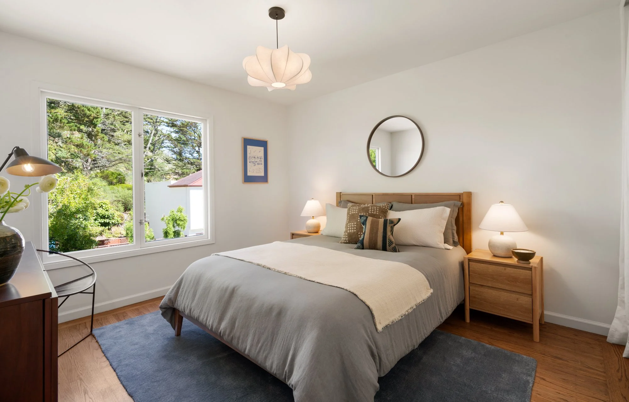 A cozy bedroom with a large bed, beige bedding, and multiple pillows. Two wooden nightstands with lamps are on each side of the bed. A round mirror hangs above the bed's headboard. A window to the left shows greenery outside, and a ceiling light fixt