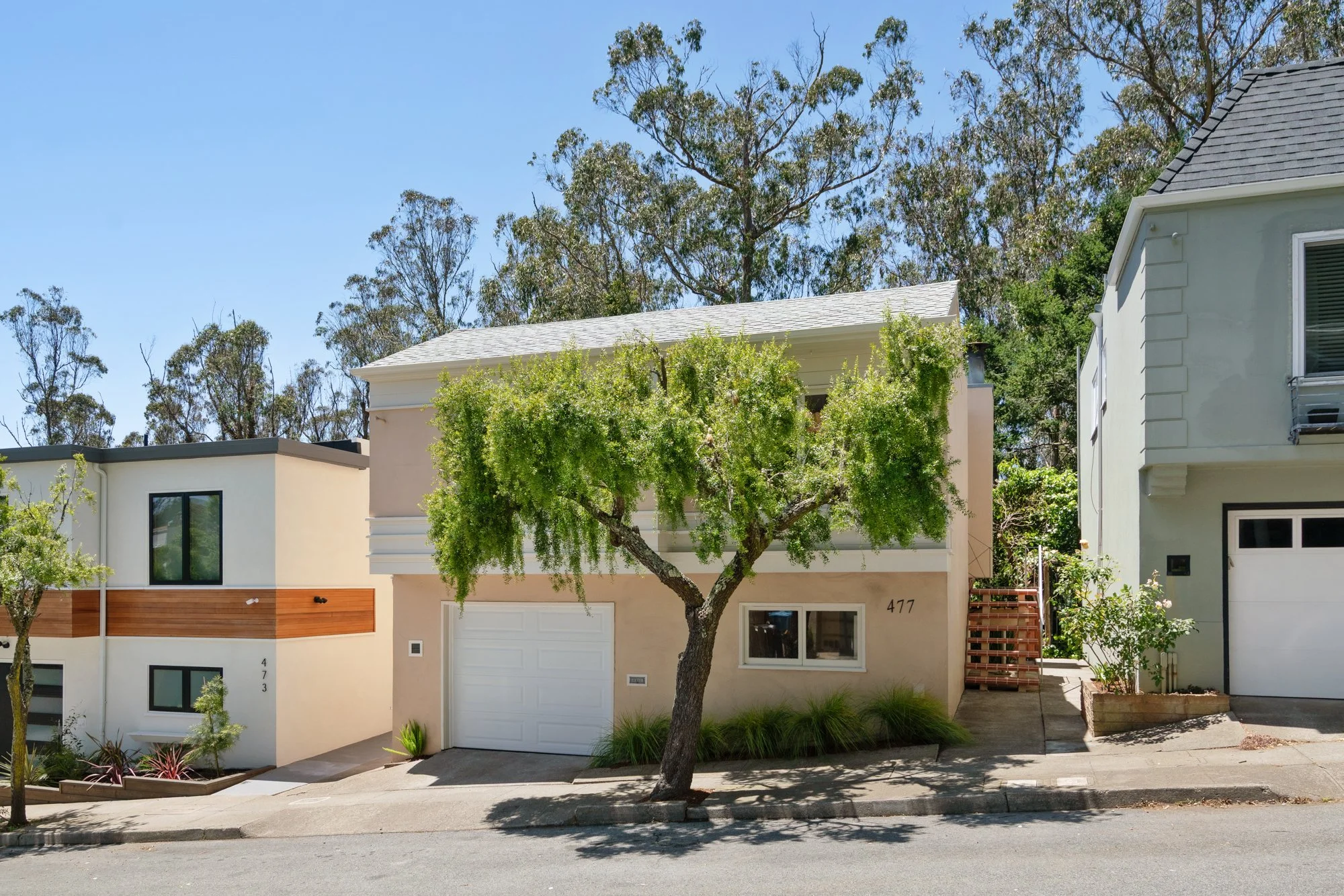 A row of modern residential houses with a tree in front, clear blue sky, and shrubbery. The house in the middle has a white garage door, greenery, and the street in the foreground.