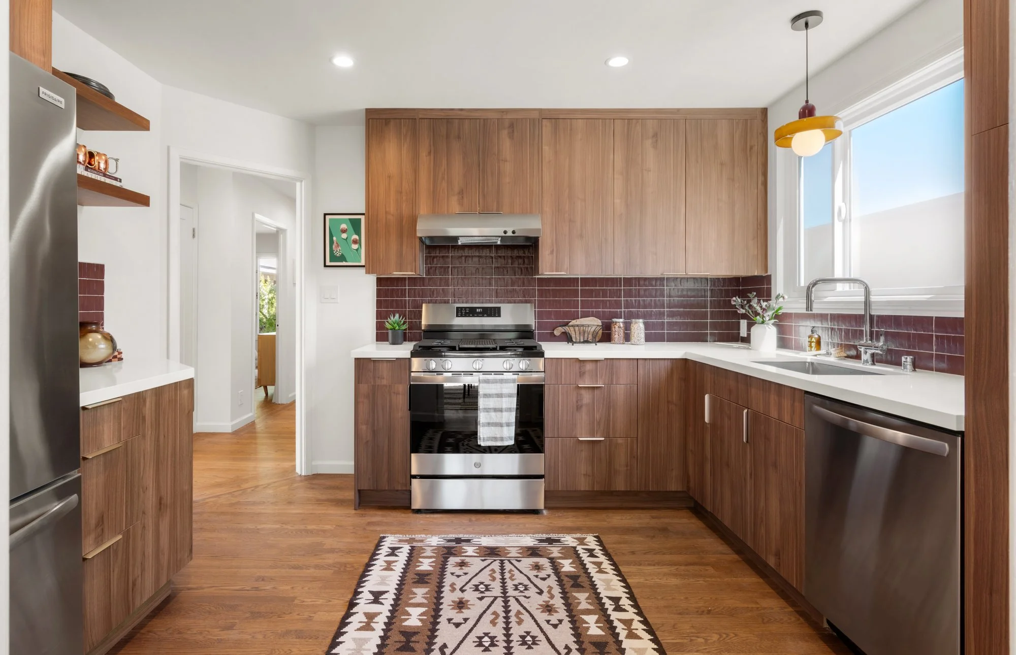 Modern kitchen with wooden cabinets, stainless steel appliances, a red tile backsplash, and a window with a view of a blue sky. A decorative rug is on the wooden floor.