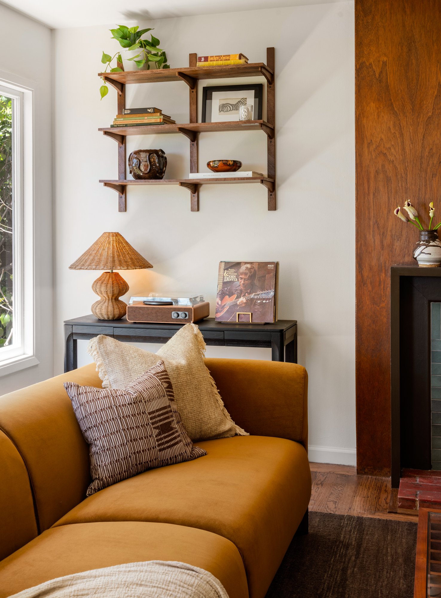 Living room corner with a mustard-colored sofa and patterned pillows, a wooden side table with a woven table lamp, wall-mounted wooden shelves with books and decorative items, a vinyl record and artwork, and a window with greenery outside.