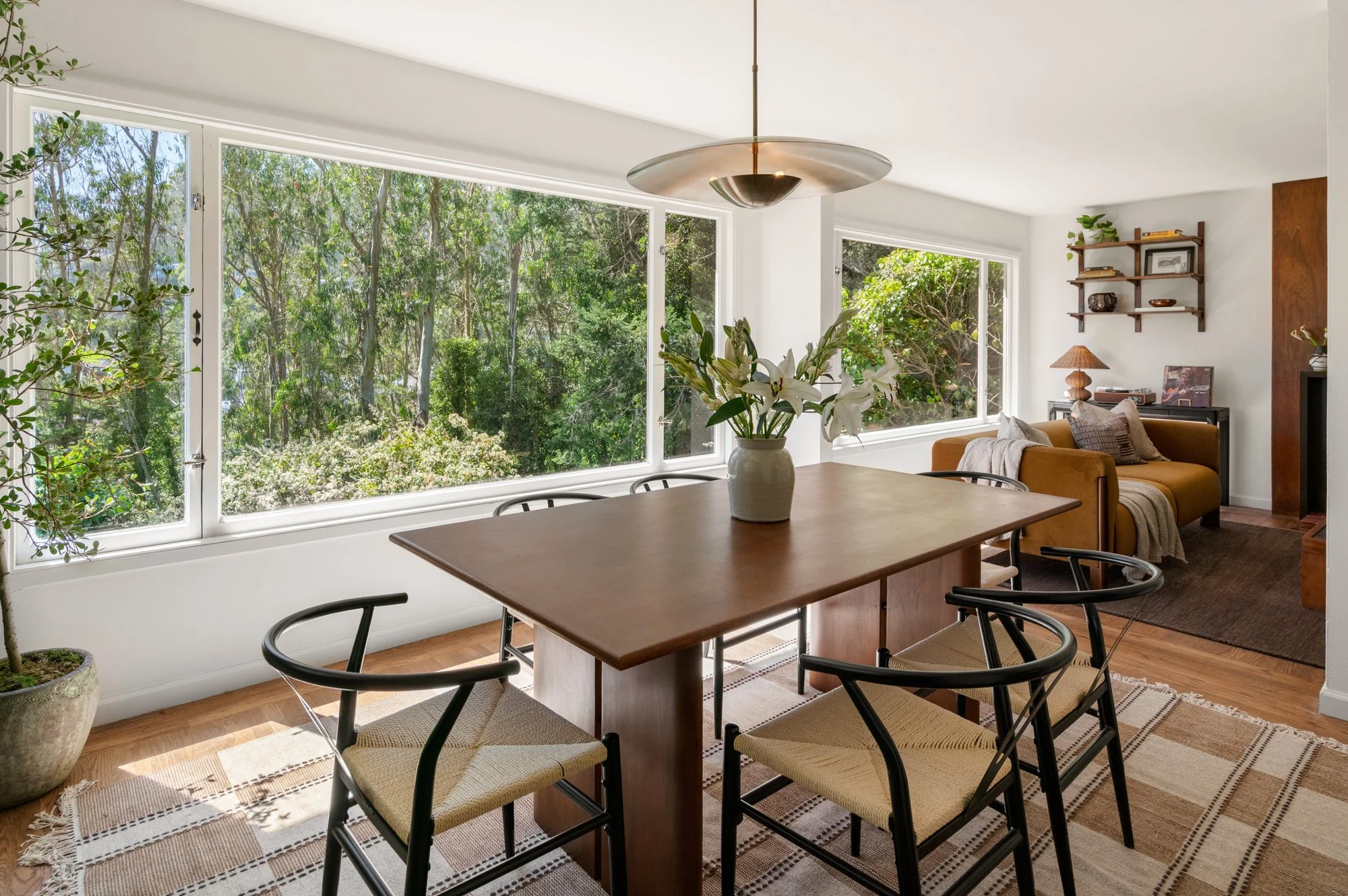Living room with large windows showing green trees outside, a wooden dining table with black chairs, a vase of white lilies, and a cozy seating area with a mustard-colored sofa and wall shelves.