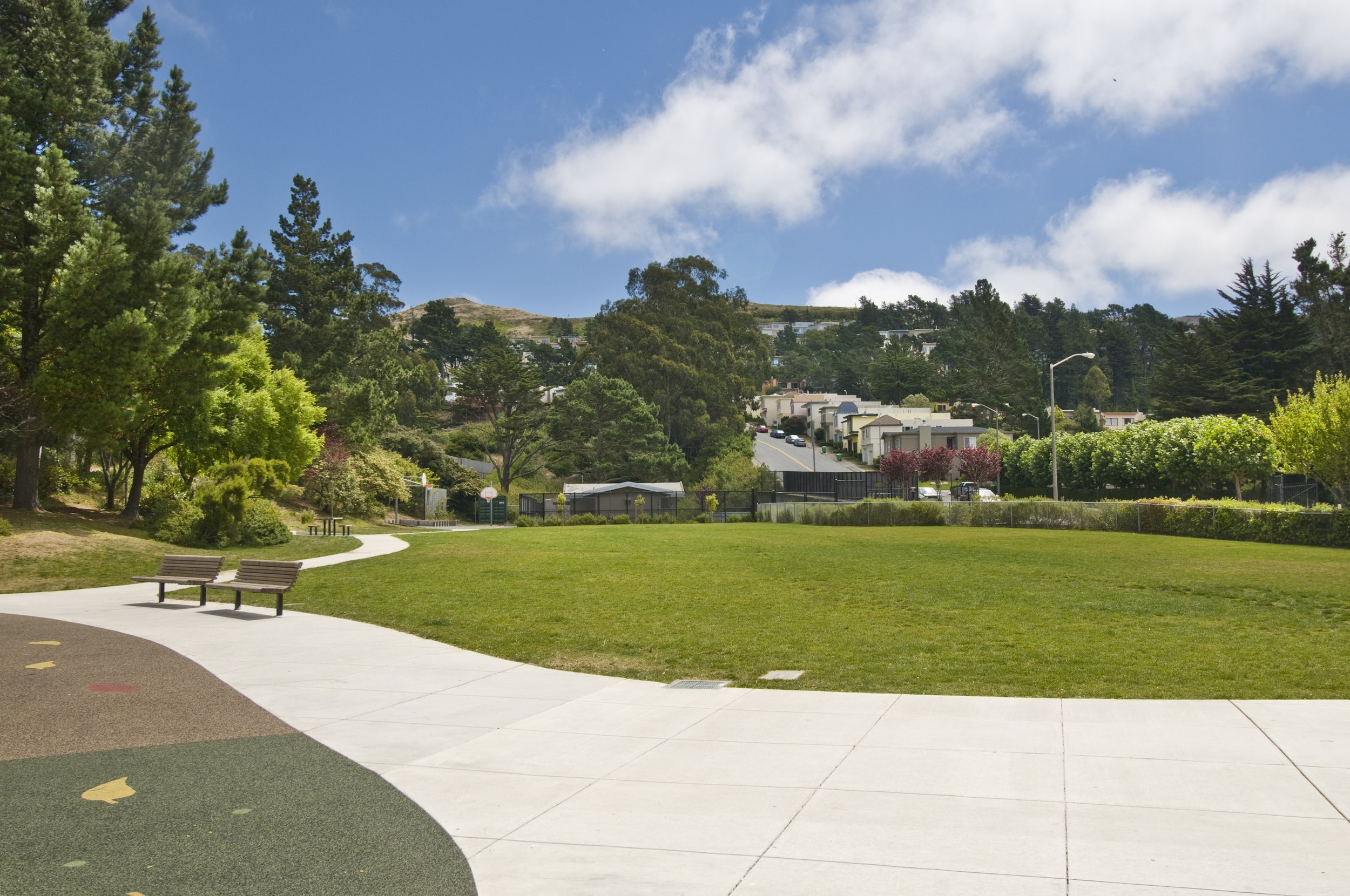 A park with green grass, trees, benches, and a walking path in the foreground. In the background, there are residential buildings on a hillside and a partly cloudy sky.