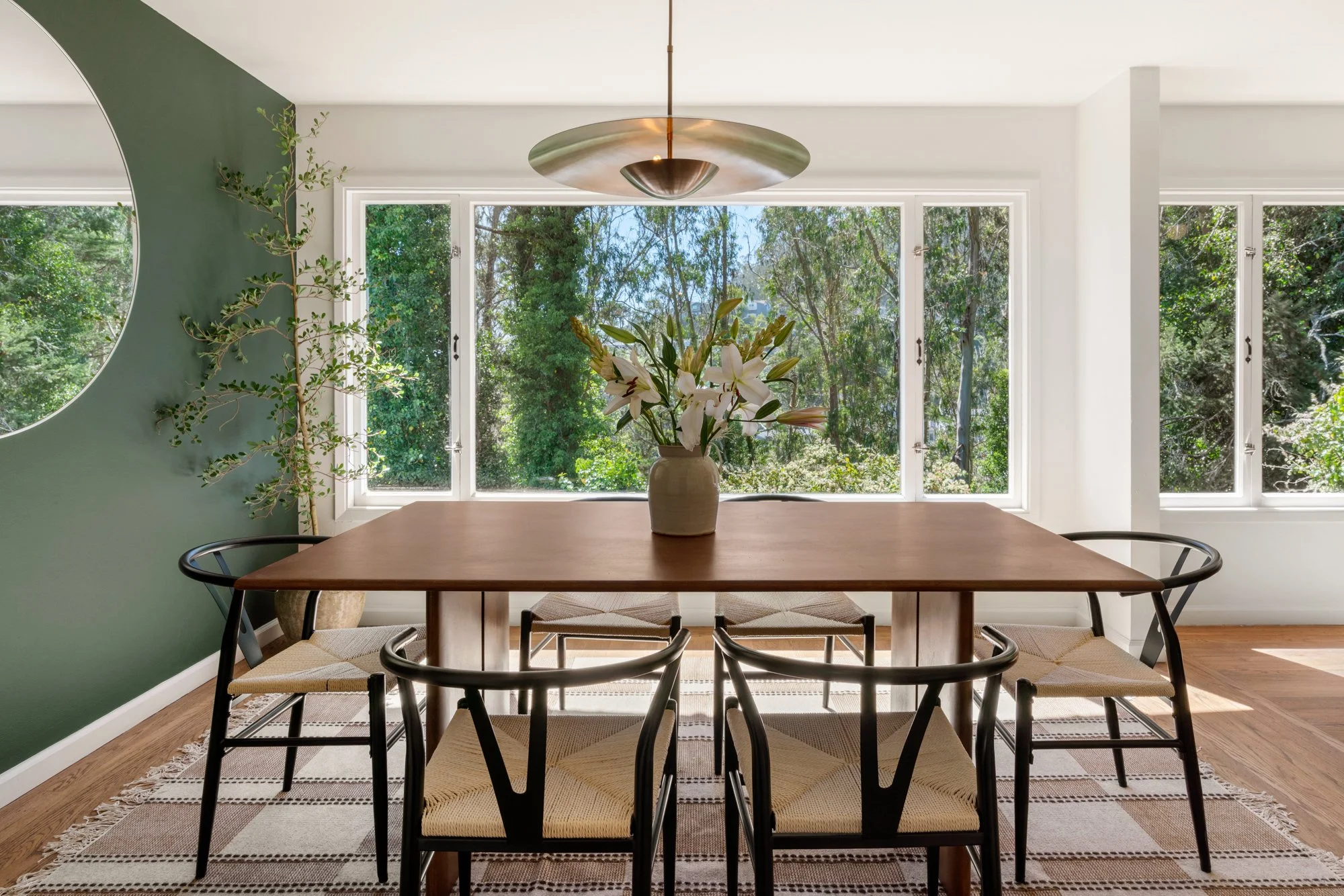 A dining room with a wooden table, black chairs with woven seats, and a patterned rug. There is a bouquet of white lilies in a beige vase on the table. The room has large windows showing green trees outside, and a modern hanging ceiling light.